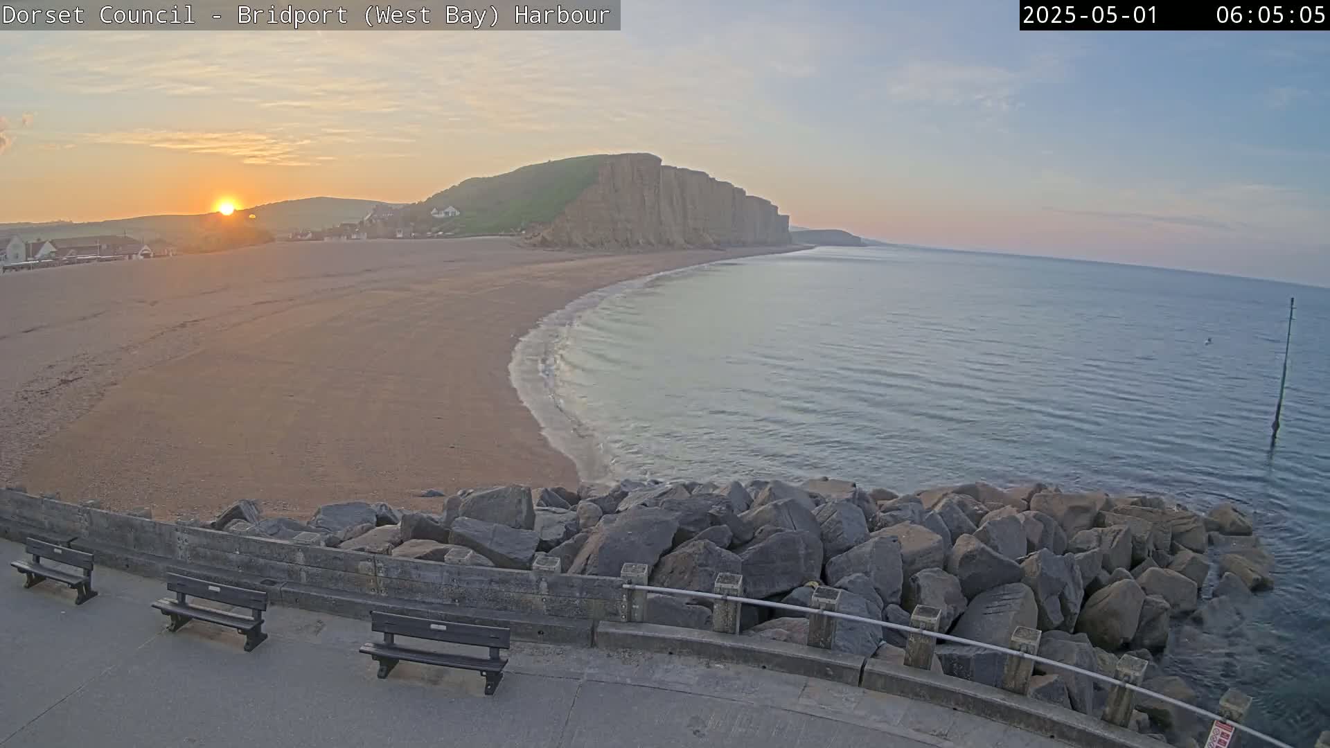 A sunrise illuminates a sandy beach curving toward a high cliff face, with calm water lapping the shore and dark rocks forming a barrier along a paved walkway.