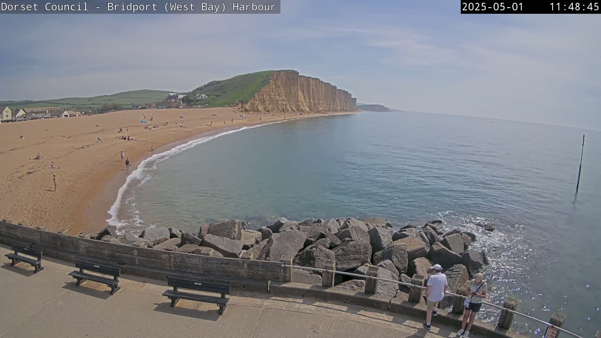 A sunny day at a beach with a sandy shore, calm water, and tall cliffs in the background, with a few people on the beach and two people near a rock wall in the foreground.