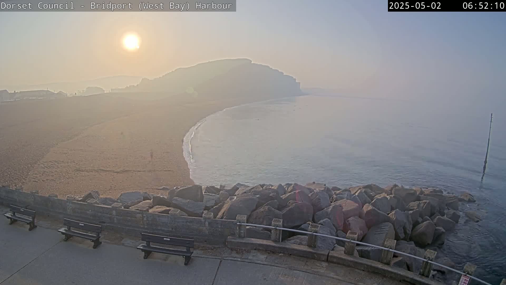 A hazy sunrise illuminates a sandy beach curving into a calm sea, with large rocks forming a barrier between the beach and a paved walkway containing benches.