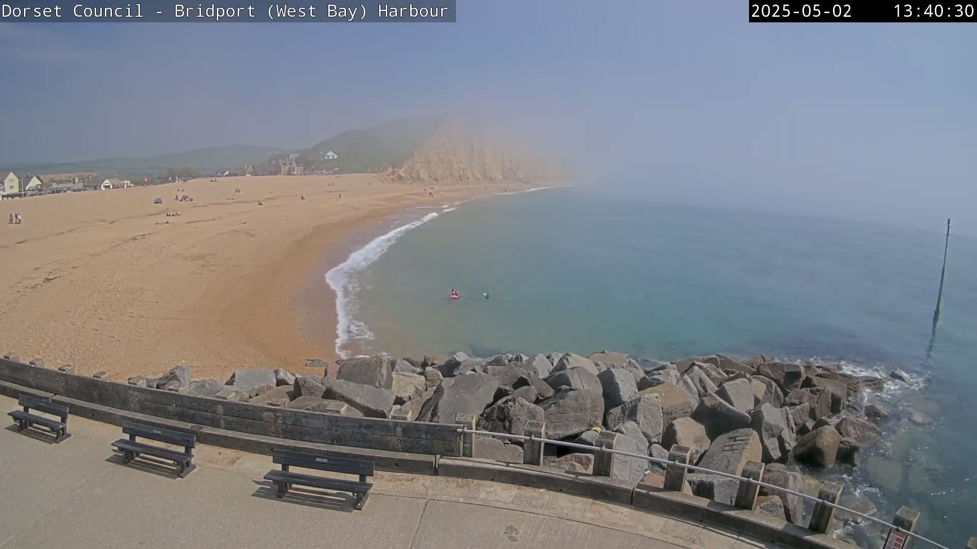 A hazy, sunny day at a sandy beach with calm, turquoise water,  backed by a cliff and featuring a rocky breakwater and several park benches.