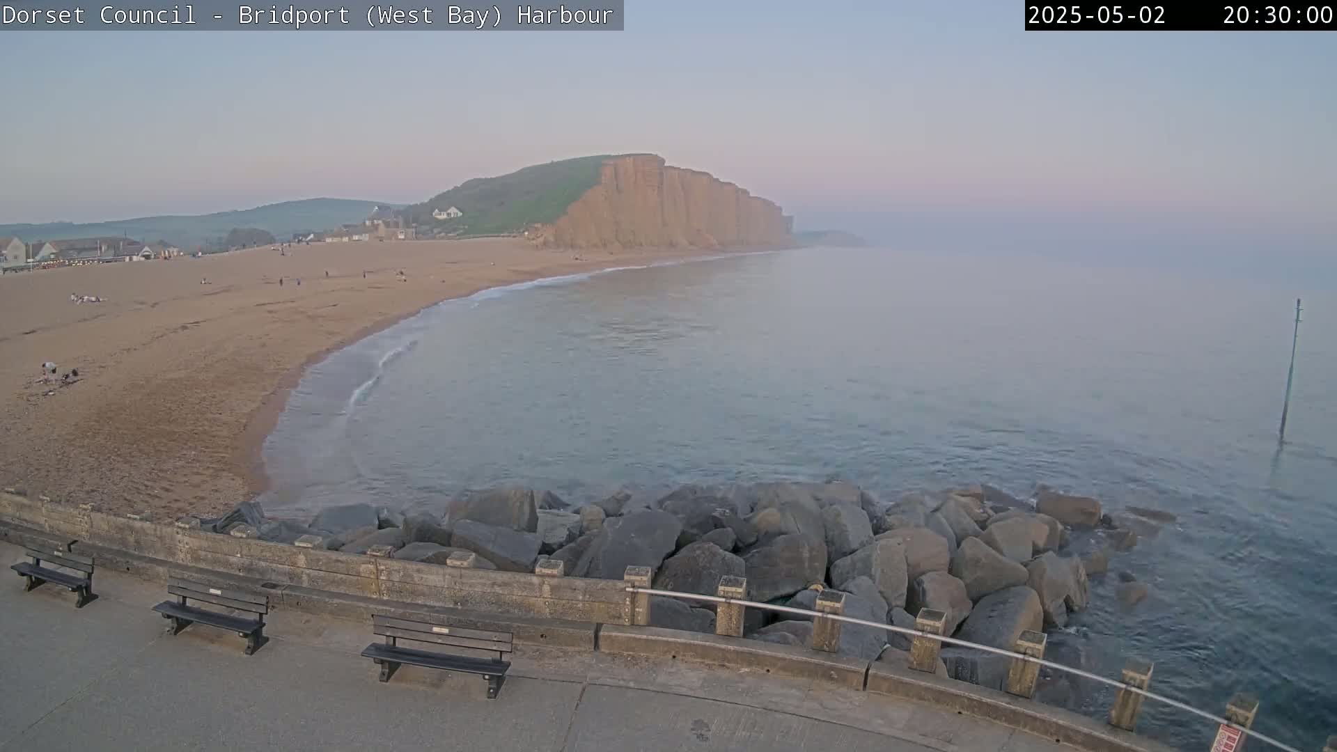 A hazy evening scene shows a sandy beach curving toward a tall, reddish cliff overlooking a calm sea, with a stone seawall and benches in the foreground.