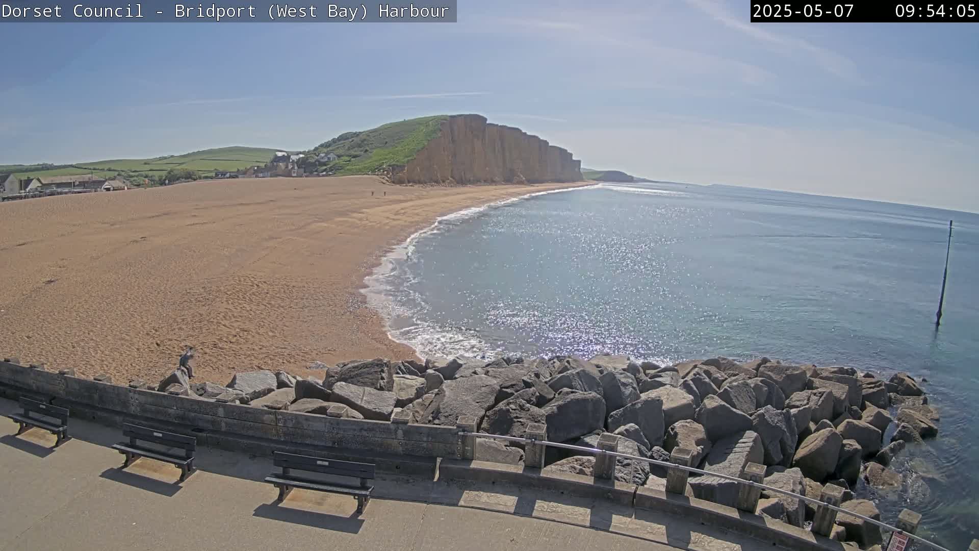 A sandy beach curves alongside a rocky coastline, meeting a calm, sunlit sea under a clear sky.