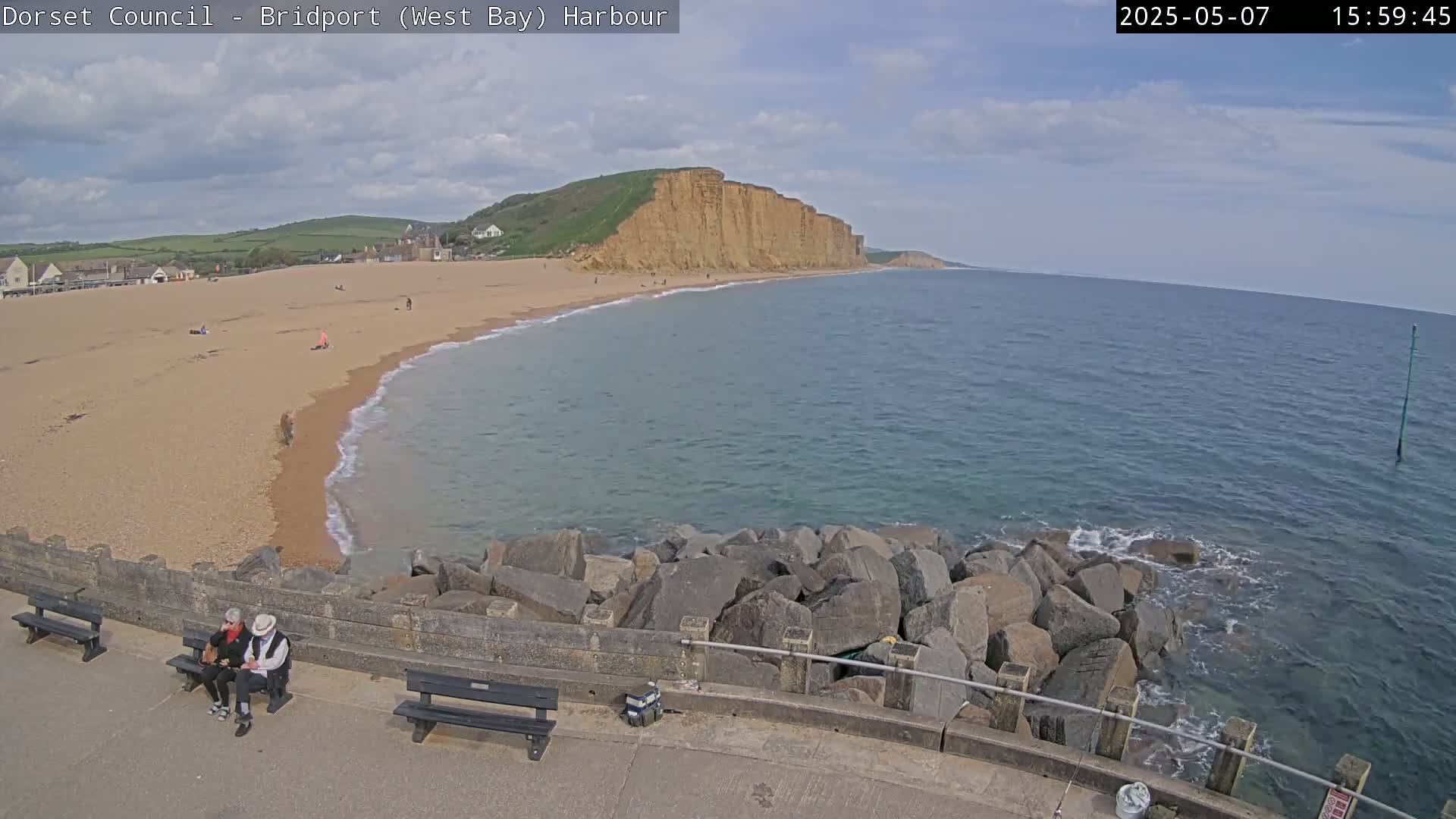 A sandy beach curves around a calm sea, meeting a rocky breakwater under partly cloudy skies; two people sit on a bench overlooking the scene.