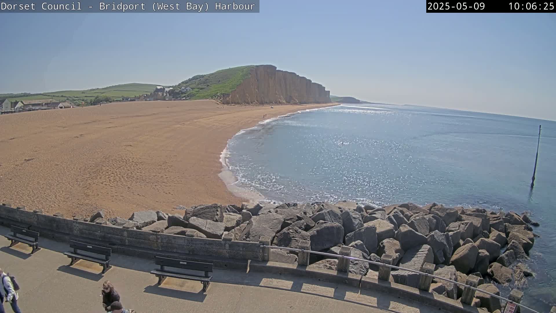 A sandy beach curves around a rocky outcropping, meeting a calm, sparkling blue ocean under a clear, sunny sky.