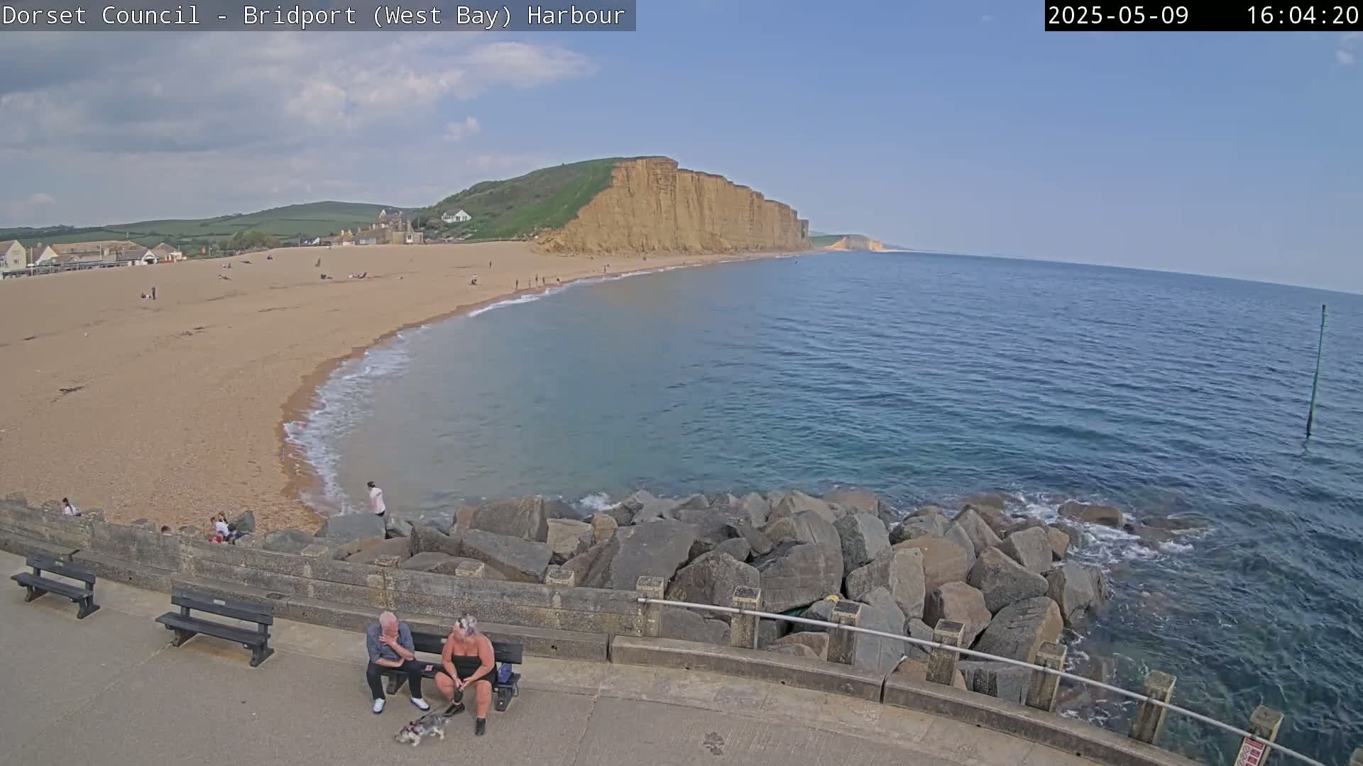 A sandy beach with people on it curves gently into a calm sea, backed by a high cliff face under a clear blue sky.