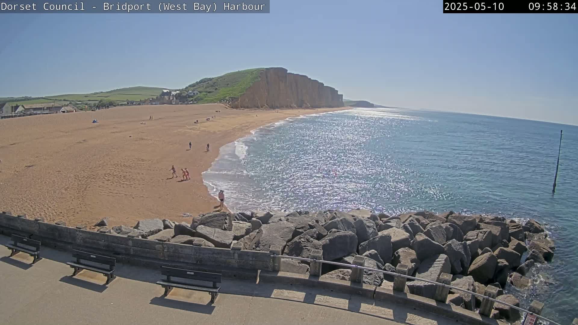 A sunny day at a sandy beach with a rocky cliff in the background, viewed from a rocky breakwater with benches.