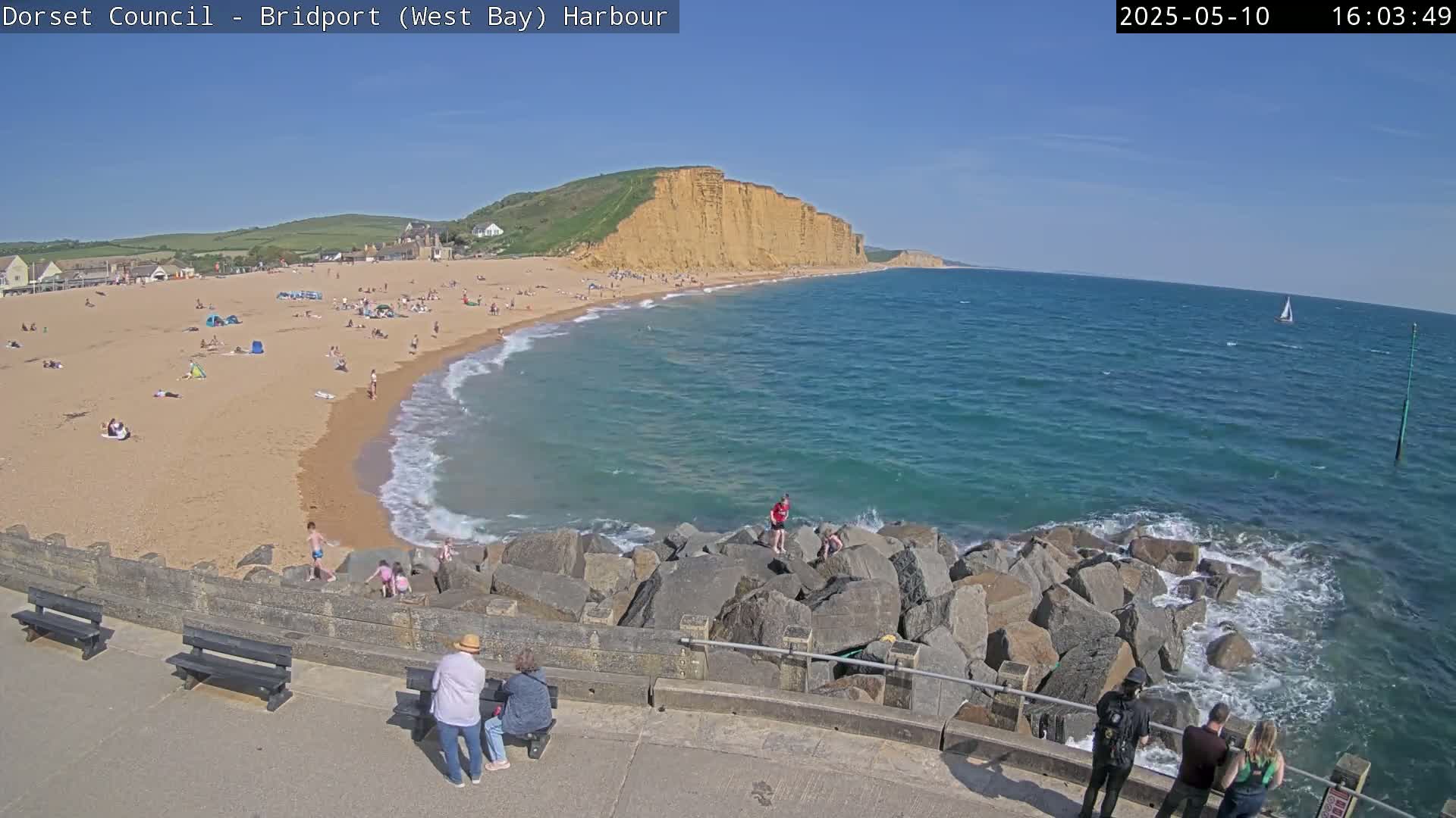 A sunny day at a beach with many people, a rocky breakwater, and a large cliff in the distance.