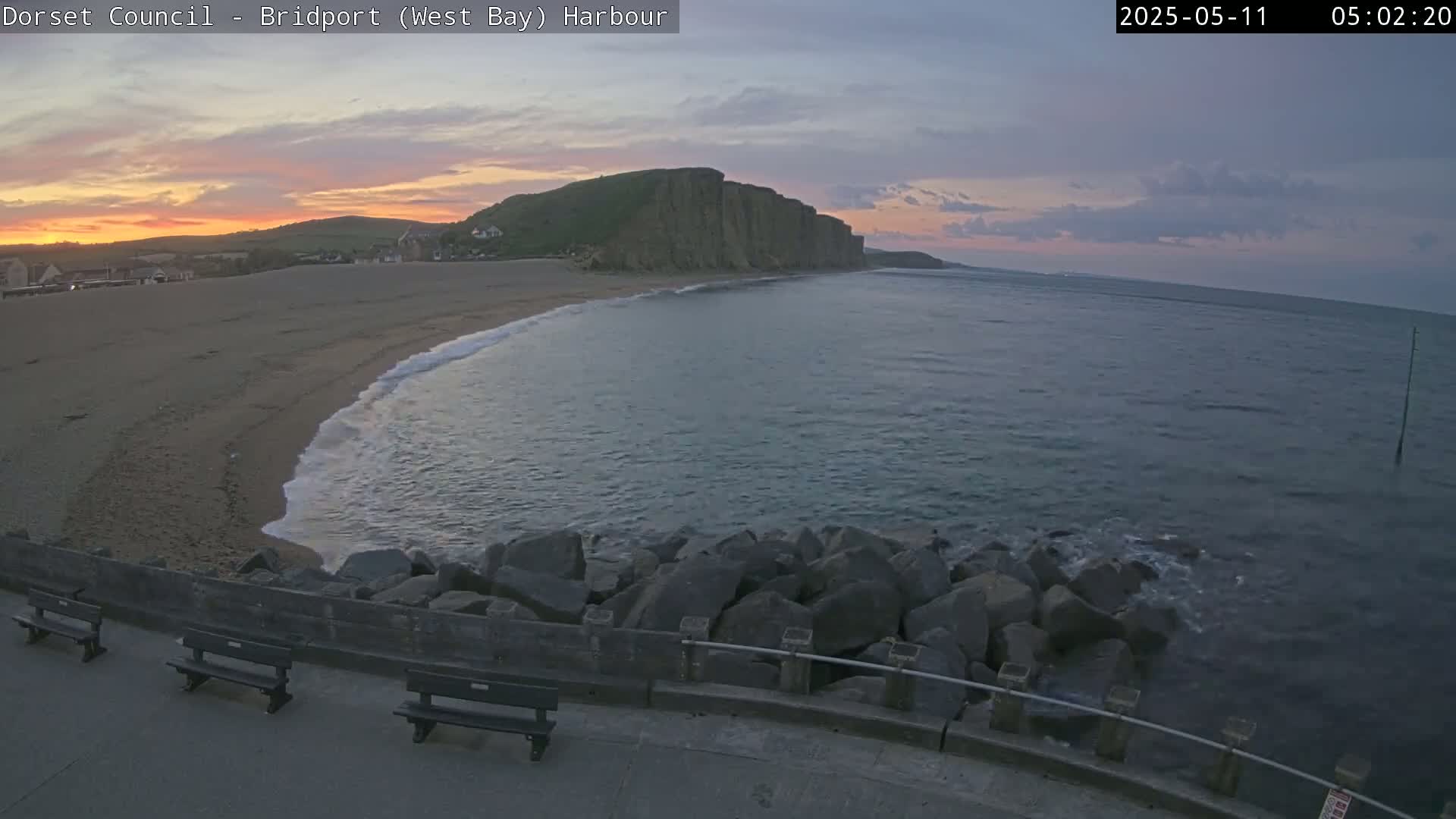 A sandy beach curves around a calm sea, with a large cliff face in the background during a sunrise.