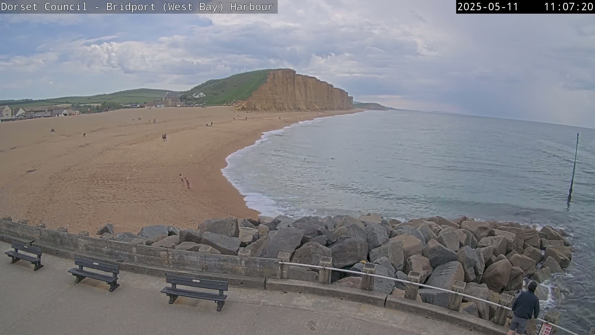 A sandy beach with a few people on it stretches beneath a cloudy sky next to a rocky cliff face, with gentle waves lapping at the shore and a rock barrier separating the beach from a paved walkway.
