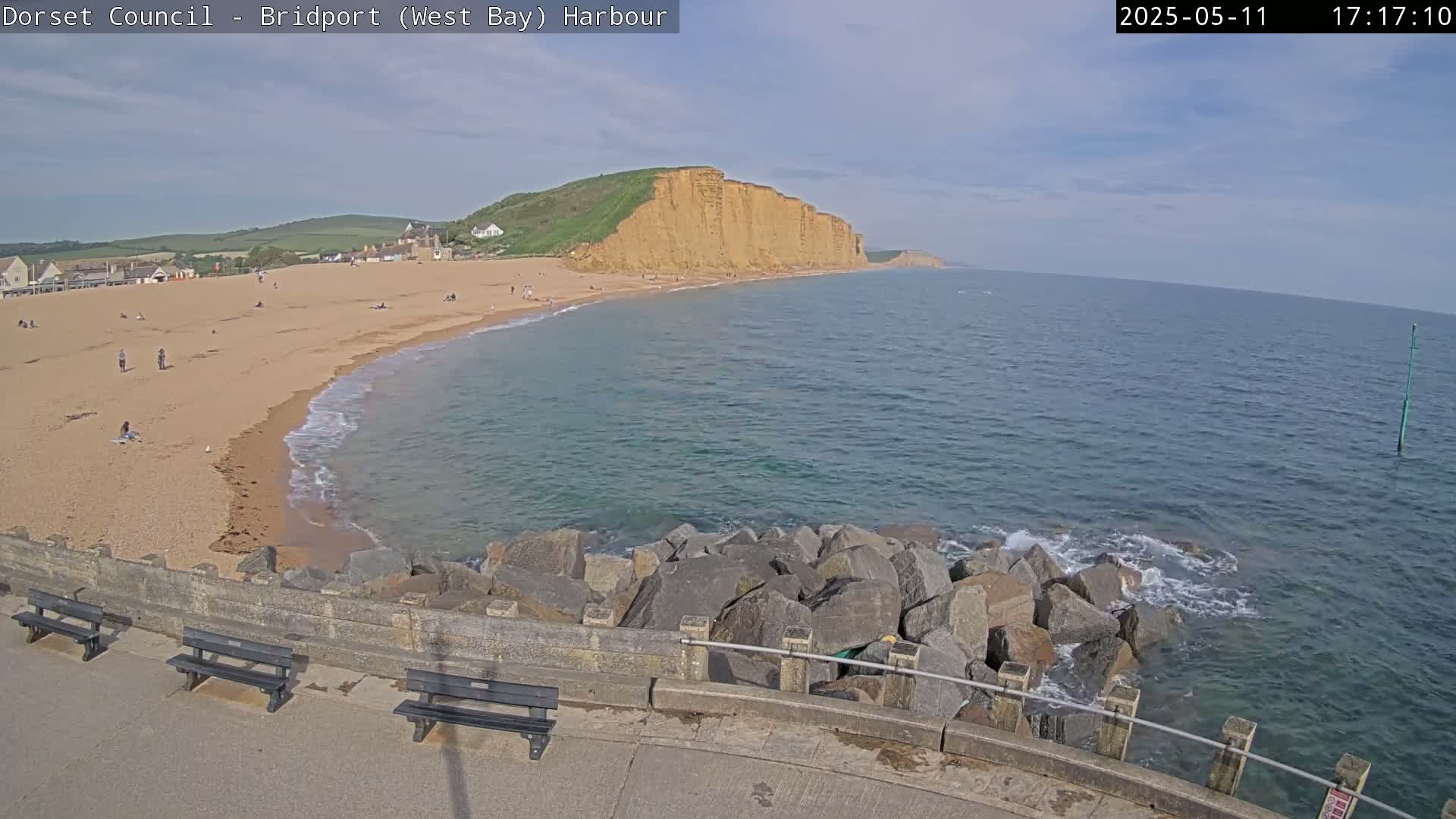 A sandy beach with people on it curves around a rocky breakwater into a calm sea under a mostly sunny sky, with tall cliffs visible in the distance.