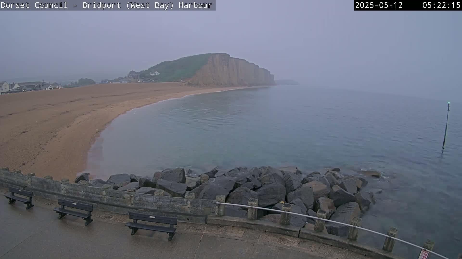 A hazy, overcast day reveals a sandy beach curving toward a rocky cliff face overlooking calm, gray water, with a low stone wall and benches in the foreground.