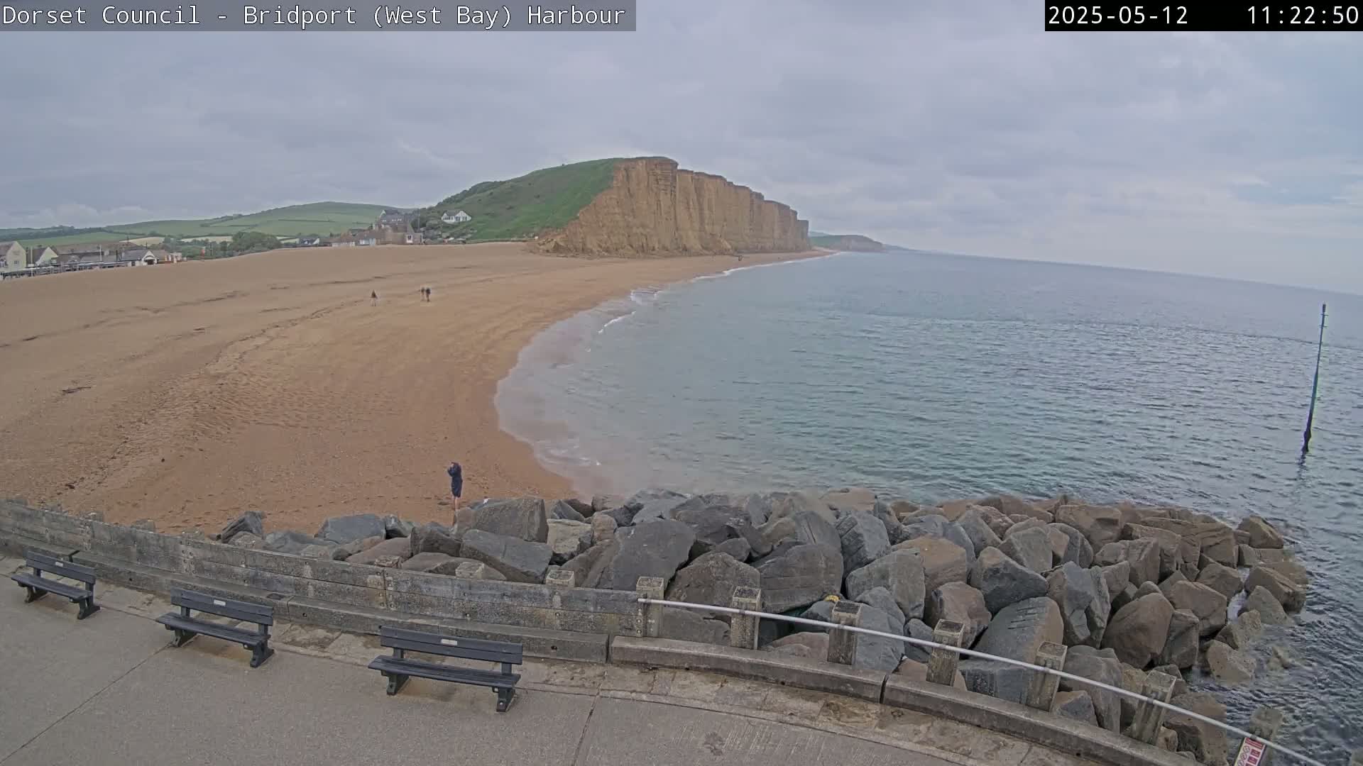 A sandy beach stretches alongside a rocky cliff face, meeting a calm, grey sea under an overcast sky.