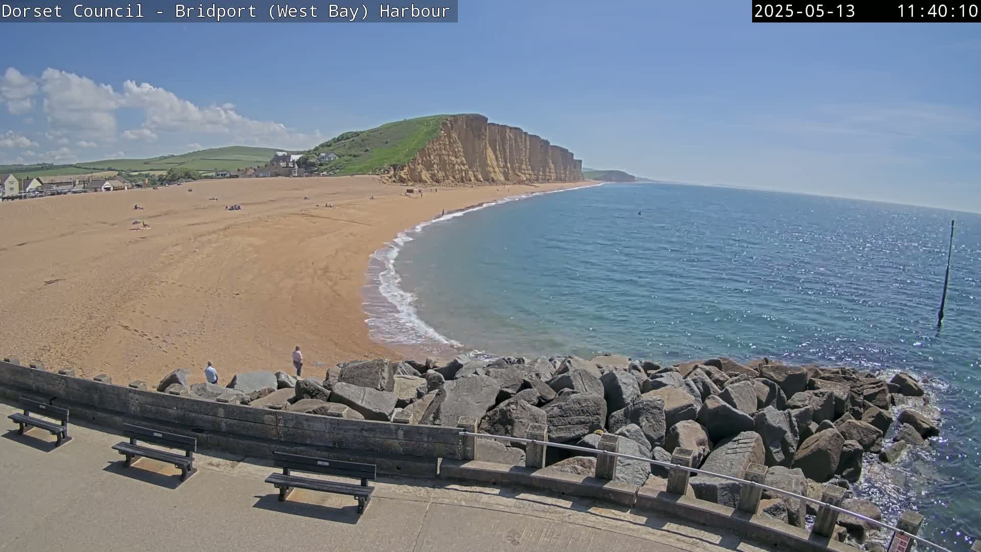 A sandy beach curves around a rocky headland into a calm, blue ocean under a mostly sunny sky.