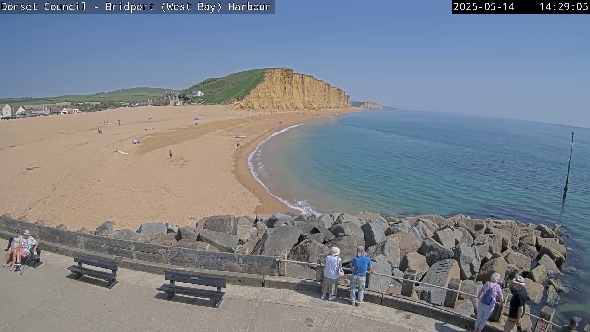 A sandy beach curves around a calm, clear ocean under a bright, sunny sky, with people scattered on the beach and a few near a rocky breakwater.