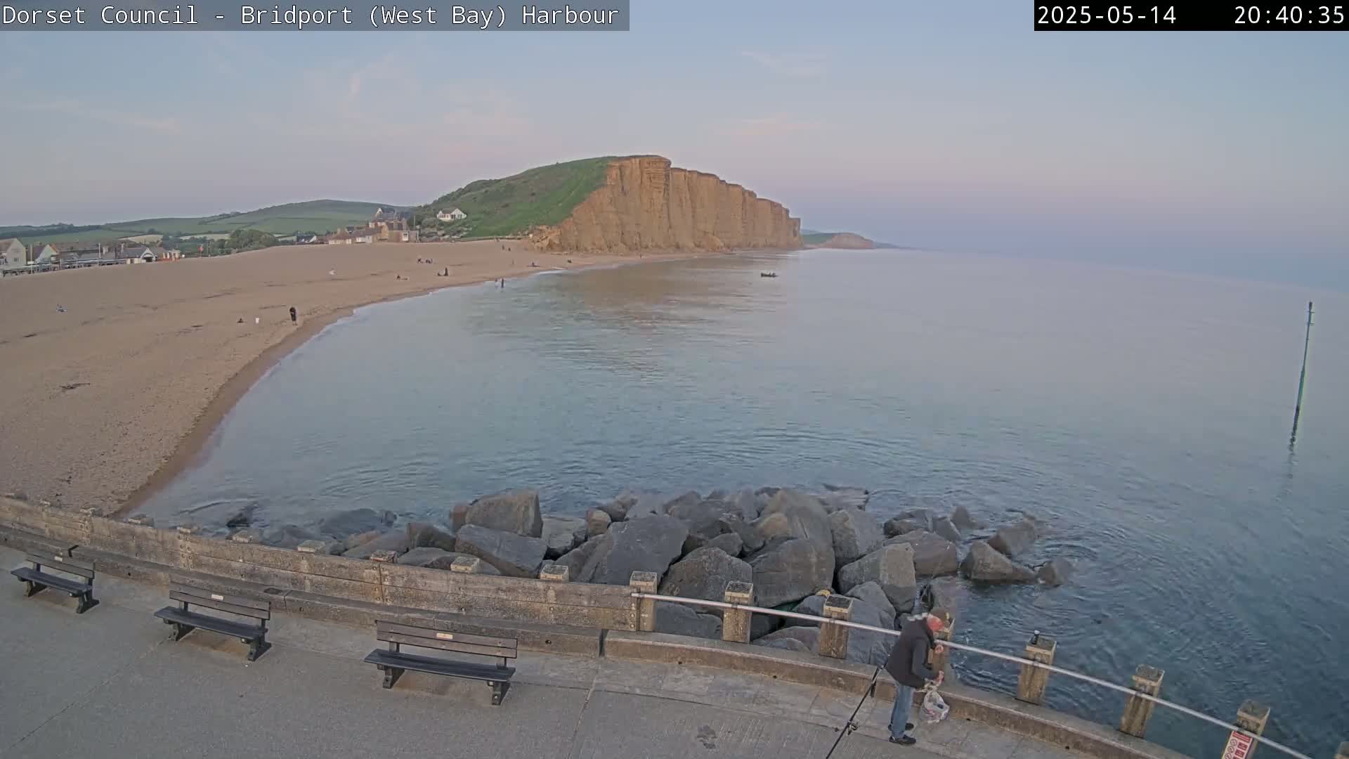 A calm, twilight ocean scene shows a sandy beach, large cliffs, a person fishing near a rocky breakwater, and several empty benches.