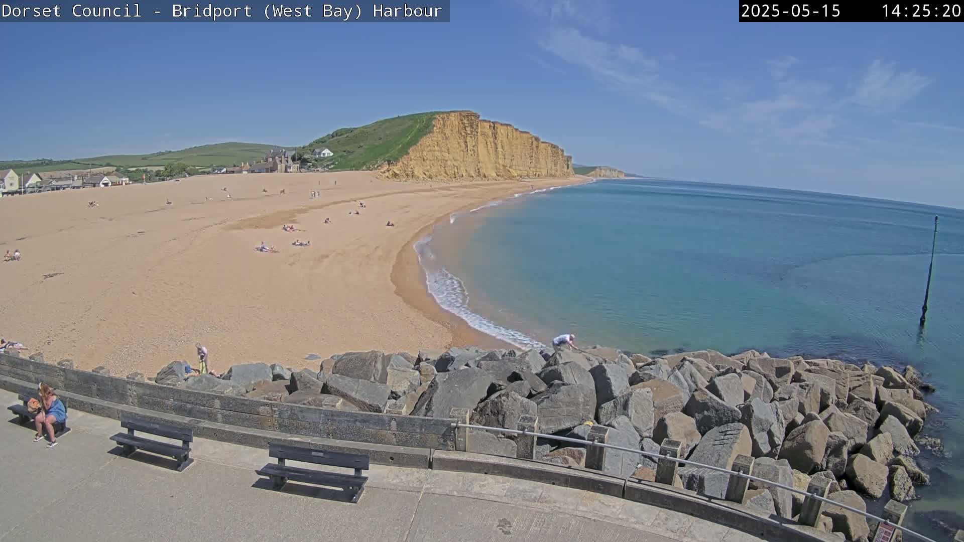 A sandy beach with people sunbathing stretches along a curved coastline under a clear blue sky, backed by high cliffs.