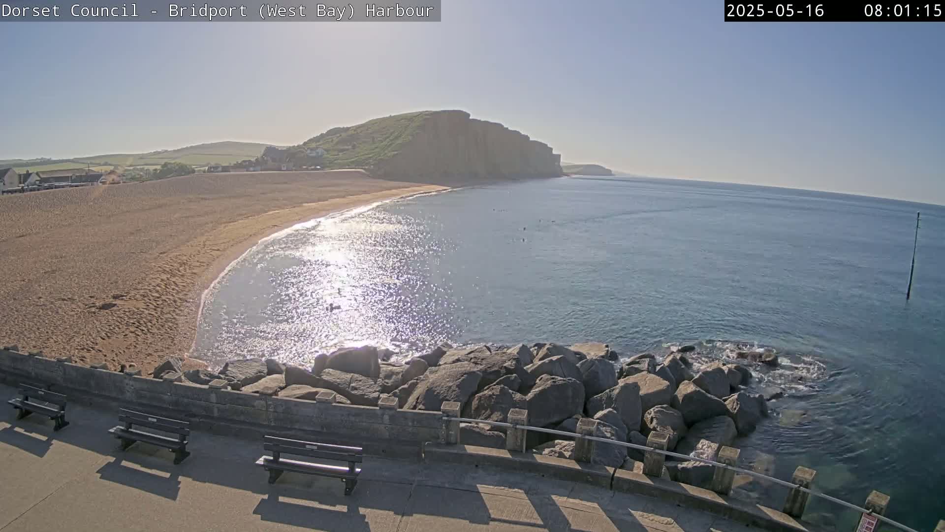 A sandy beach curves into a calm, sunlit ocean, backed by a high cliff, with several benches situated on a stone wall overlooking the water.
