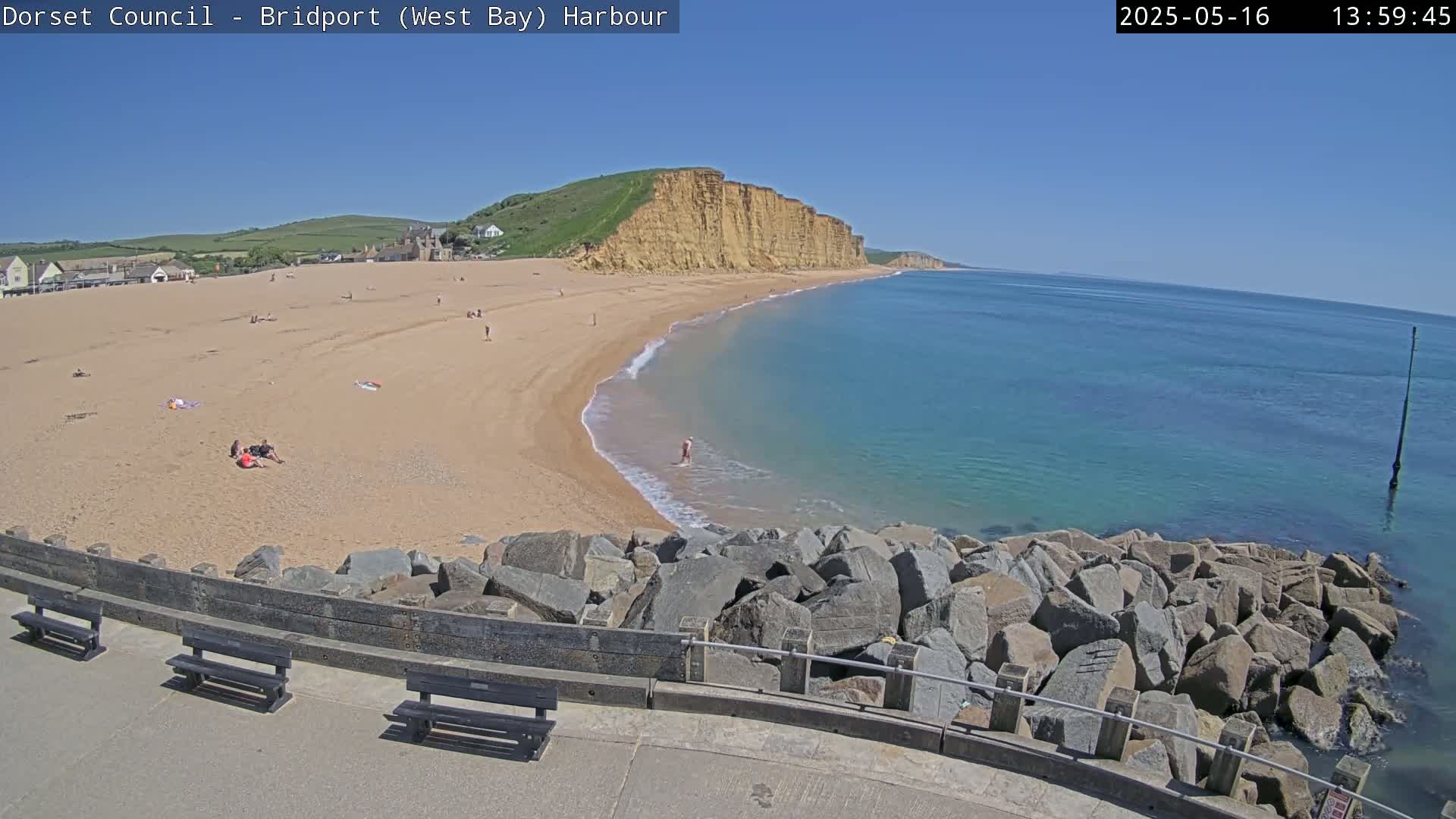 A sandy beach with people sunbathing and swimming, backed by high cliffs, stretches alongside a calm, turquoise sea under a clear blue sky.