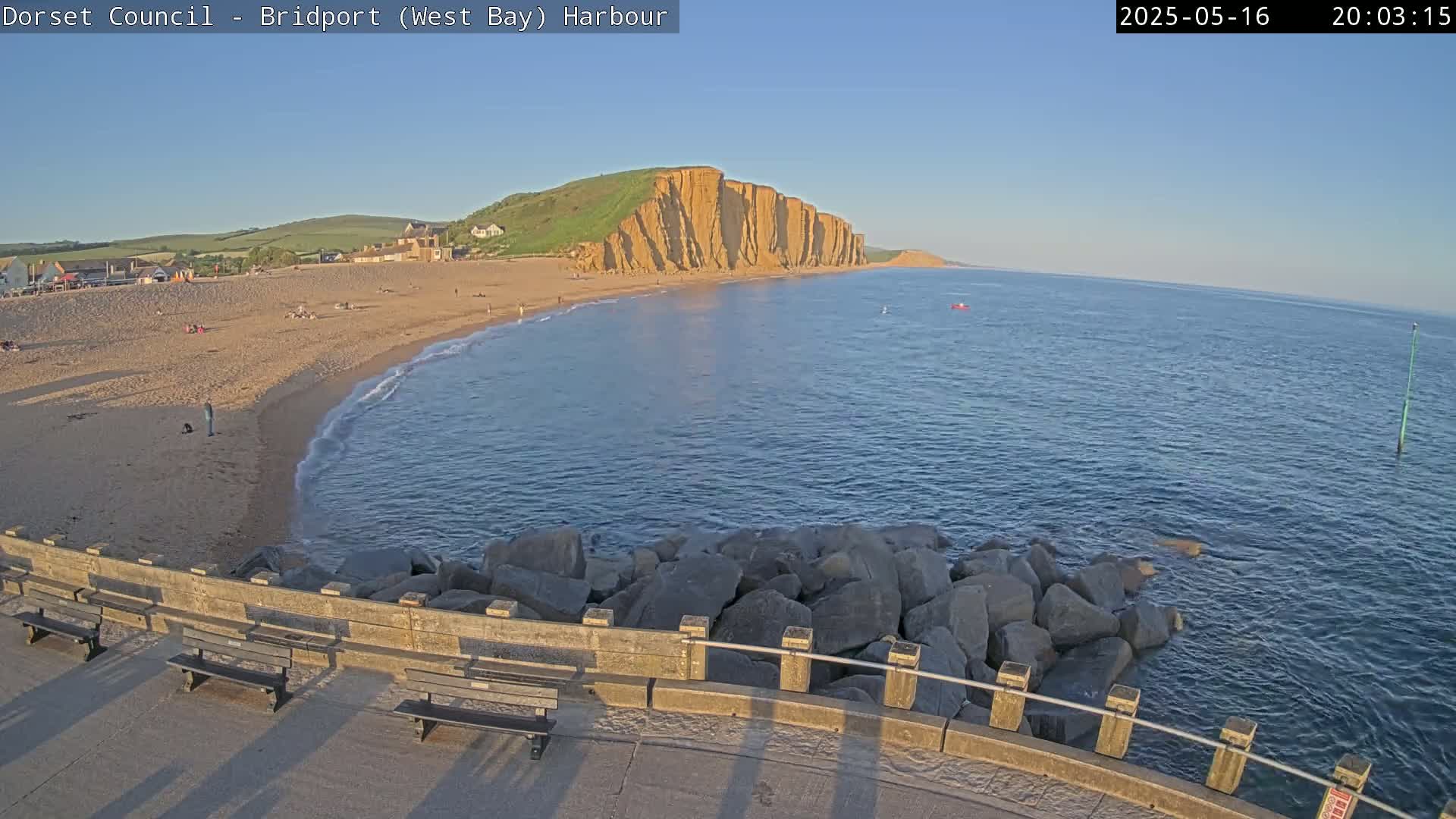 A sandy beach with a few people on it curves gently into a calm sea, under a clear evening sky, and is bordered by a rocky breakwater with benches.