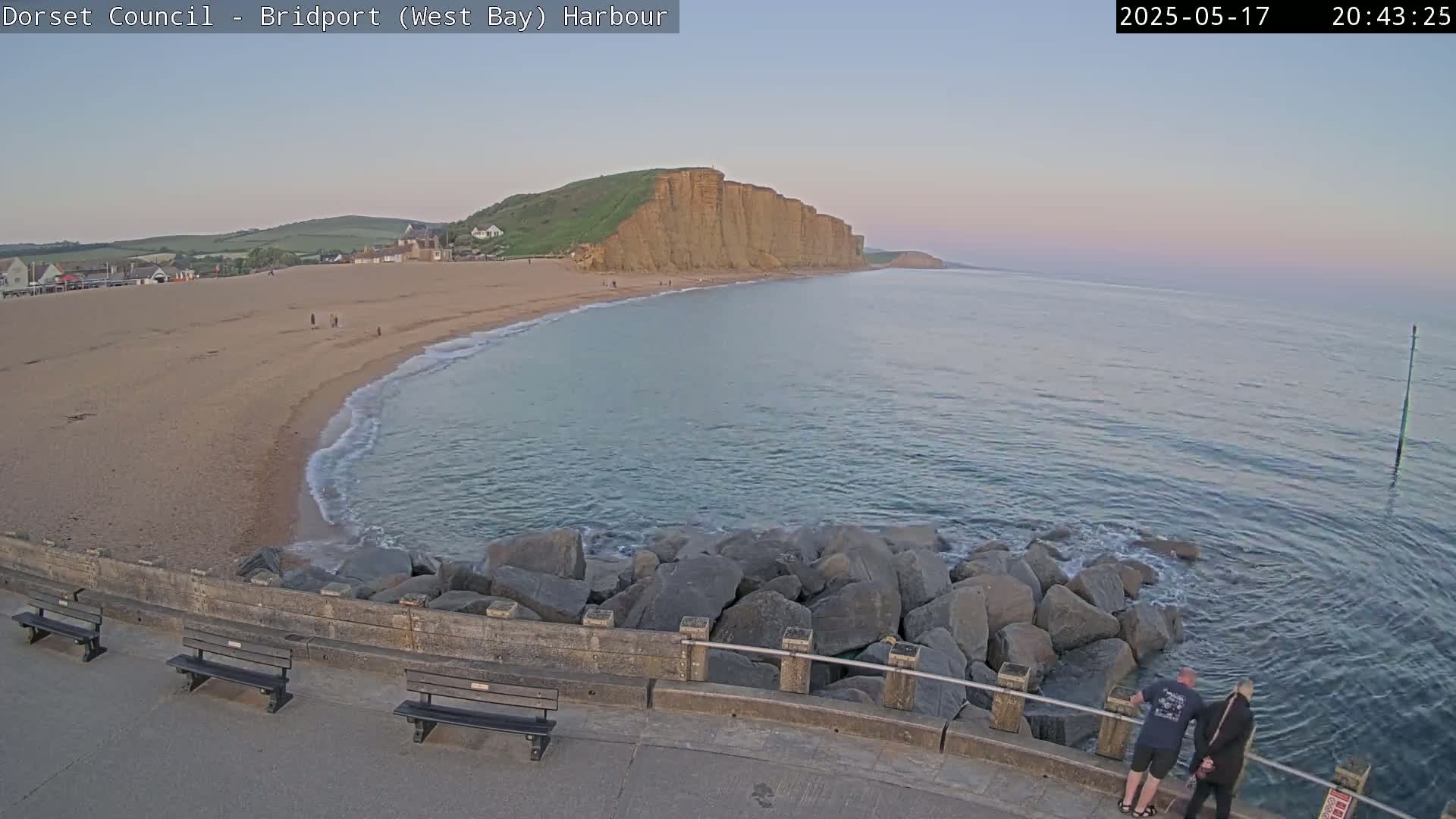 A sandy beach curves around a calm, blue-green sea at twilight, with large rocks forming a barrier and a few people standing near them.