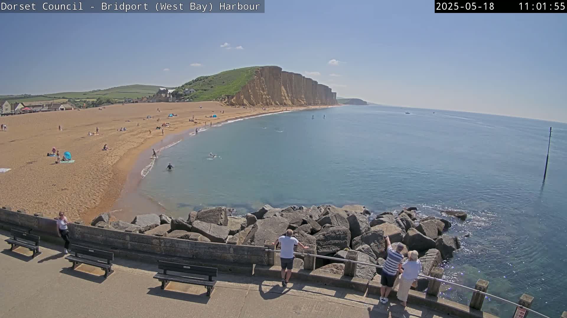 A sunny day at a sandy beach with people swimming and sunbathing, overlooking a calm ocean and towering cliffs in the distance.