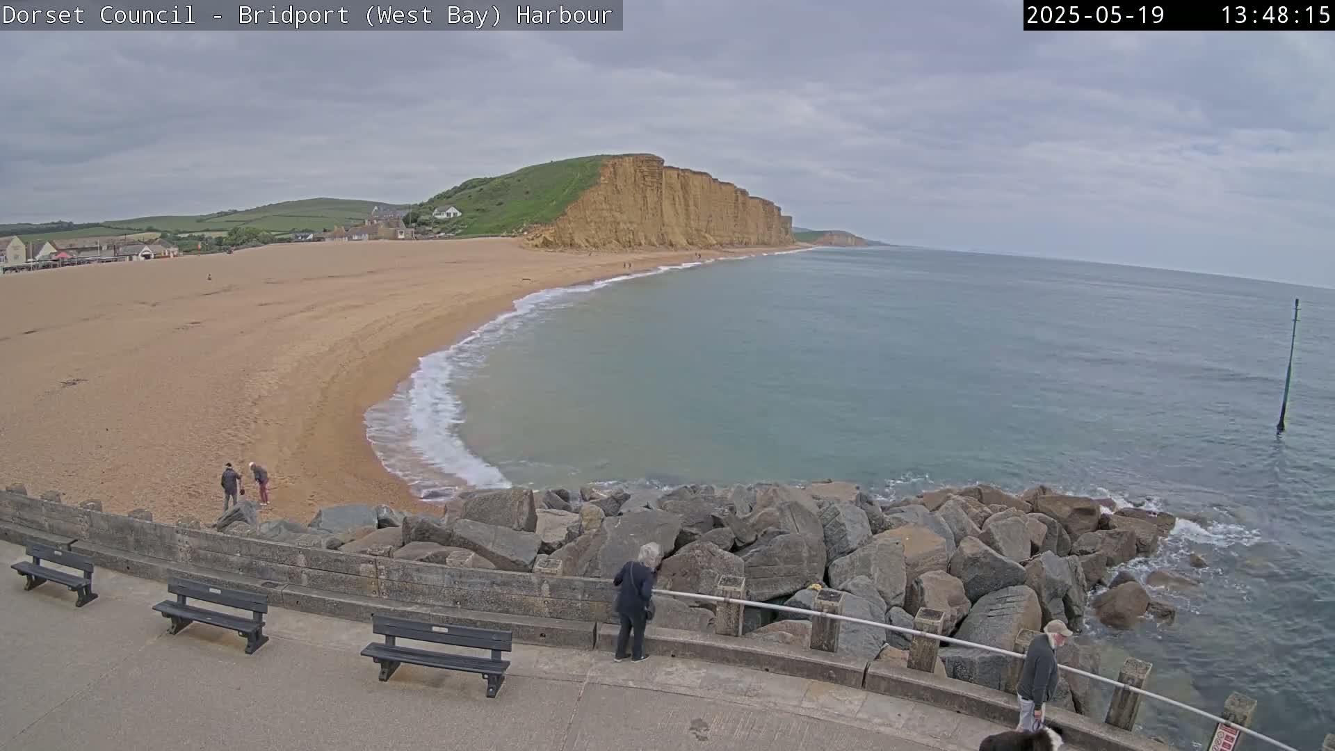 A sandy beach curves around a rocky harbor, meeting a calm sea under a cloudy sky, with a few people visible on the beach and near the water.