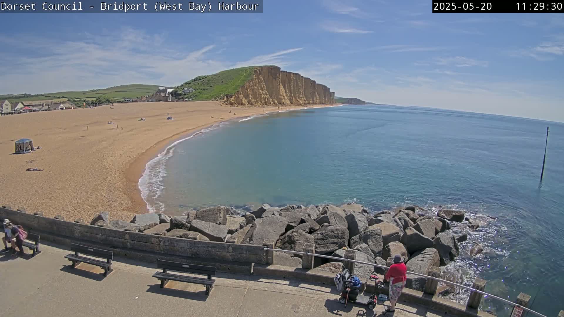 A sandy beach with a few people on it curves around a rocky breakwater into a calm, blue ocean, backed by high, tan cliffs under a mostly sunny sky.