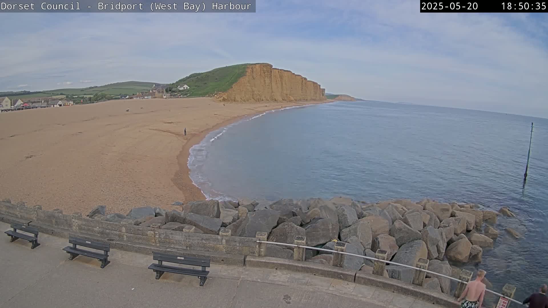A sandy beach curves around a rocky cove, meeting a calm sea under a partly cloudy sky.