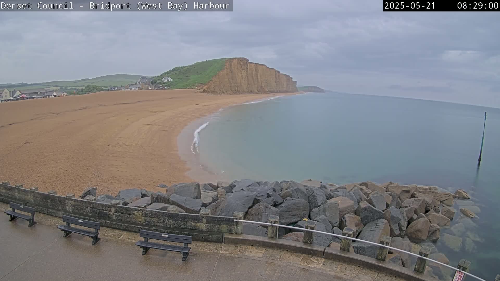 A sandy beach curves around a rocky shoreline, meeting a calm, pale-blue sea under an overcast sky.