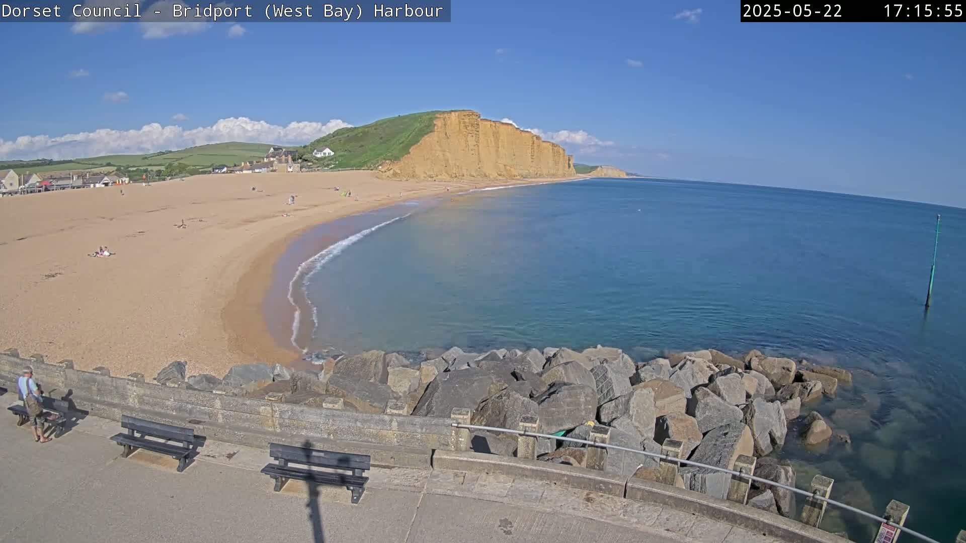 A sandy beach curves gently into a calm sea under a mostly sunny sky, backed by tall, golden cliffs.