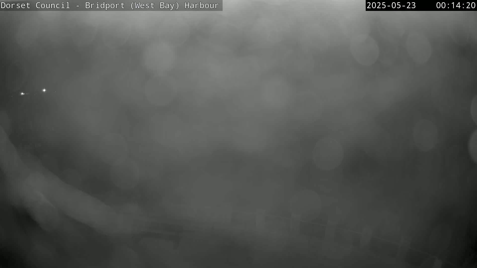 A blurry, dark gray image shows a foggy or misty outdoor scene at night, with indistinct shapes and a few faint lights visible in the distance.