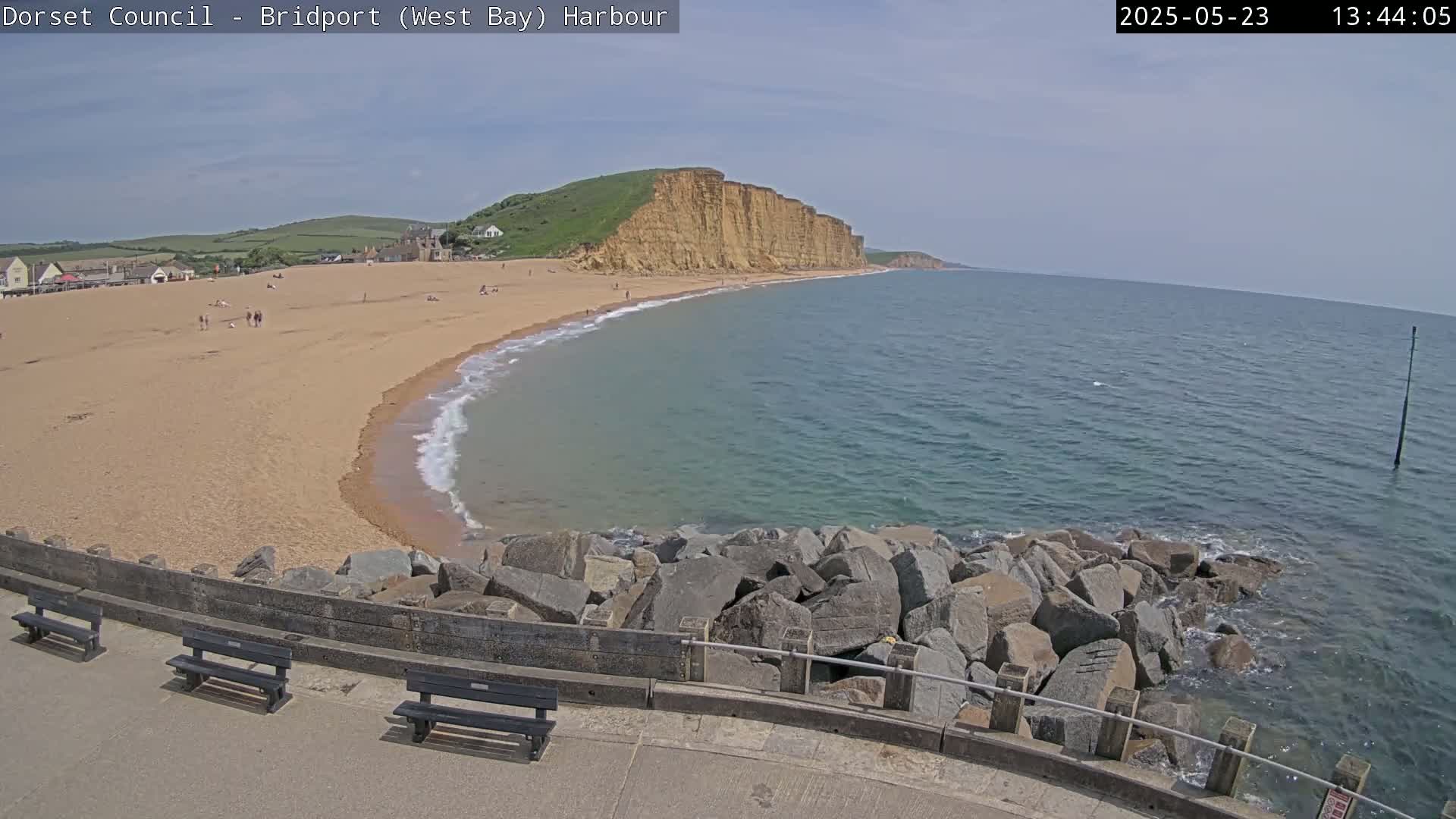 A sandy beach with people on it curves gently around a rocky headland, backed by green hills under a mostly clear sky, with a calm sea lapping at the shore.