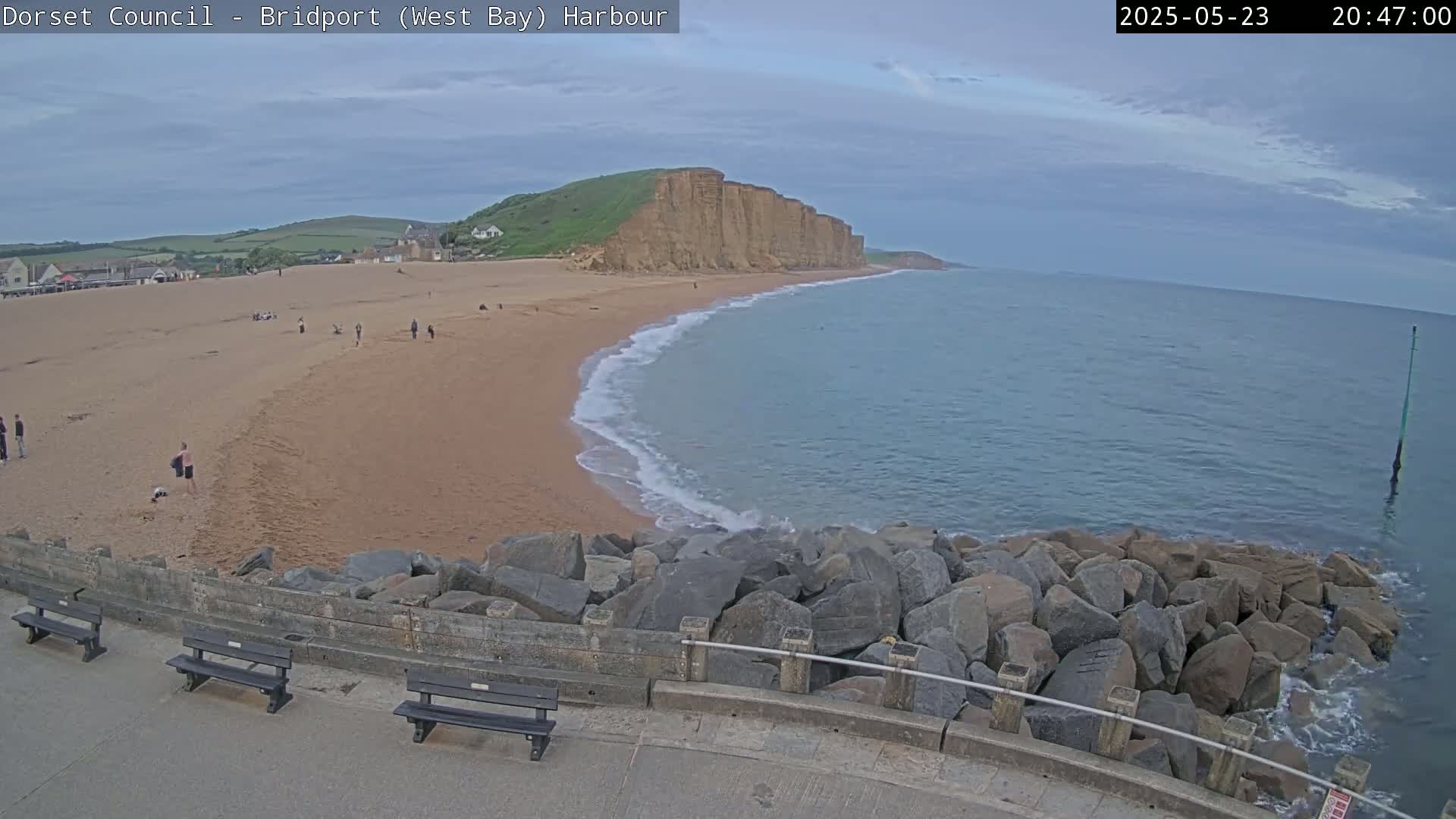 A sandy beach with a few people on it curves around to meet a calm sea under a cloudy sky, with large rocks forming a barrier between the beach and a paved area with benches.
