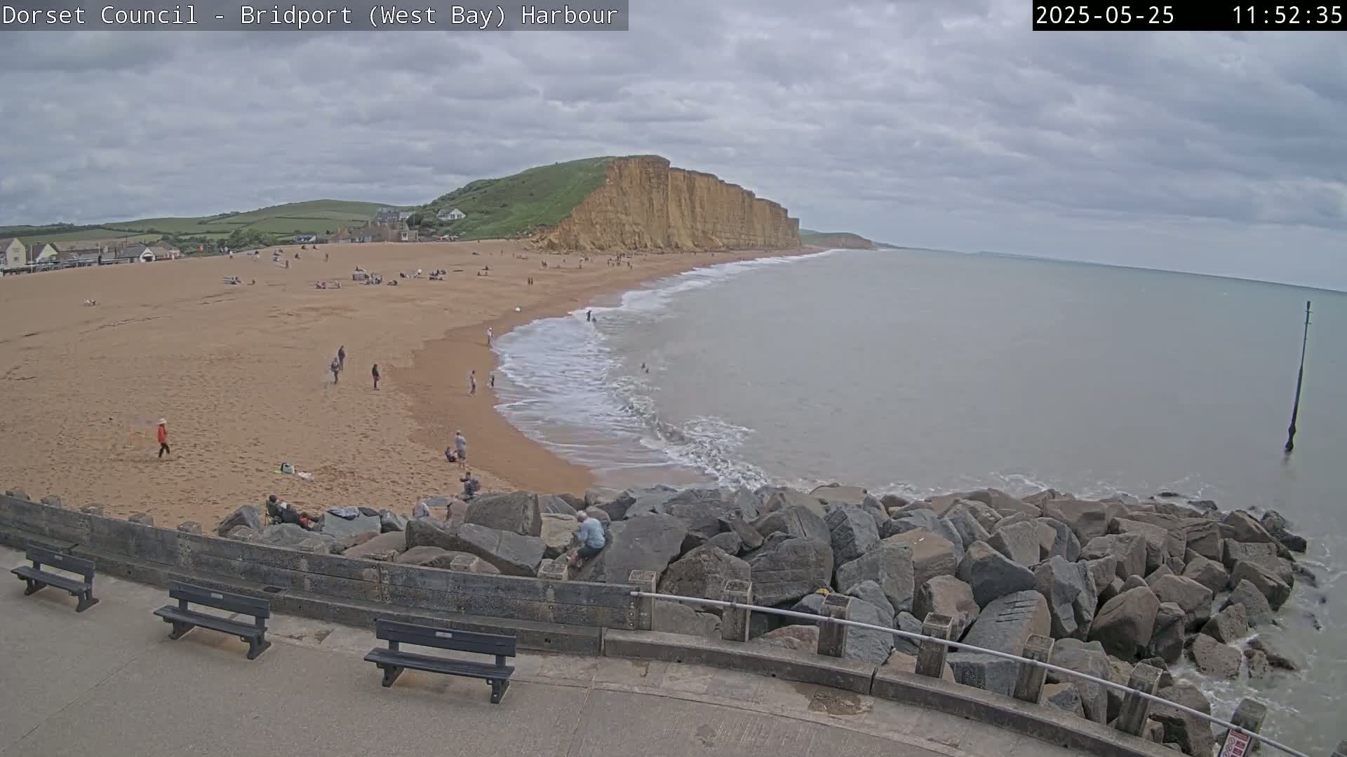 A sandy beach with people on it, bordered by a rocky seawall and overlooking a calm sea under a cloudy sky, with a large cliff in the background.