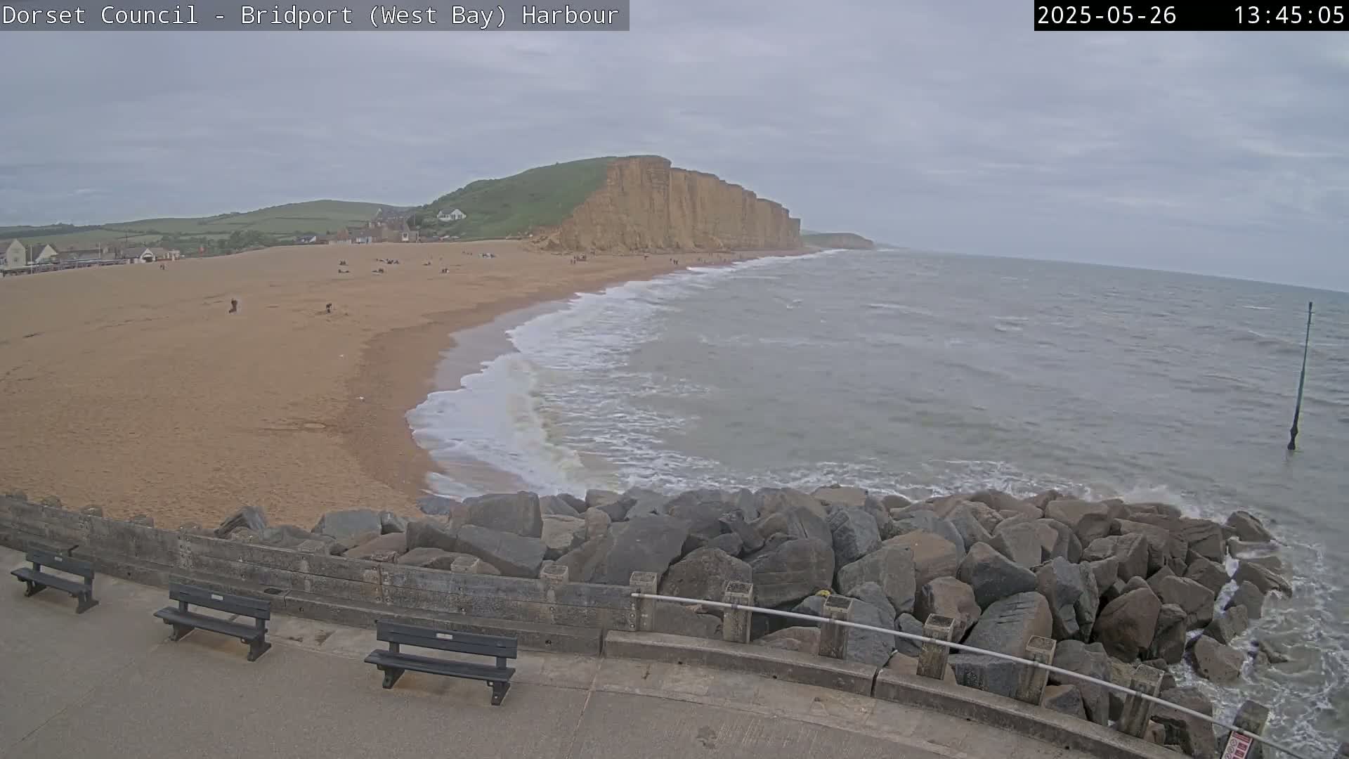 A sandy beach stretches beneath an overcast sky toward a high, cliff-faced headland, with gentle waves lapping the shore and a rocky breakwater in the foreground.