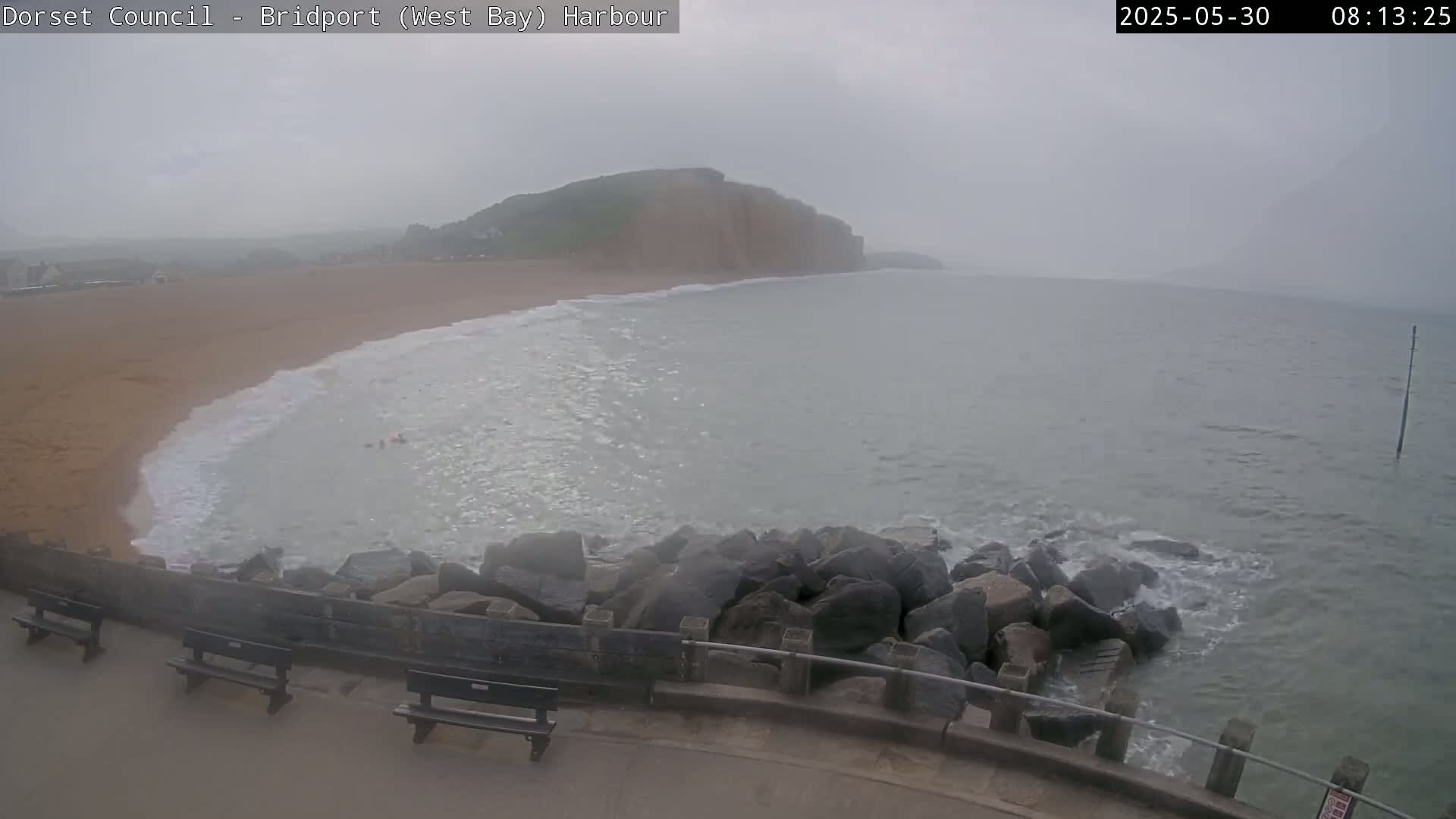 A hazy, overcast day reveals a sandy beach meeting a calm sea with small waves breaking against a rocky outcrop; several park benches sit nearby.