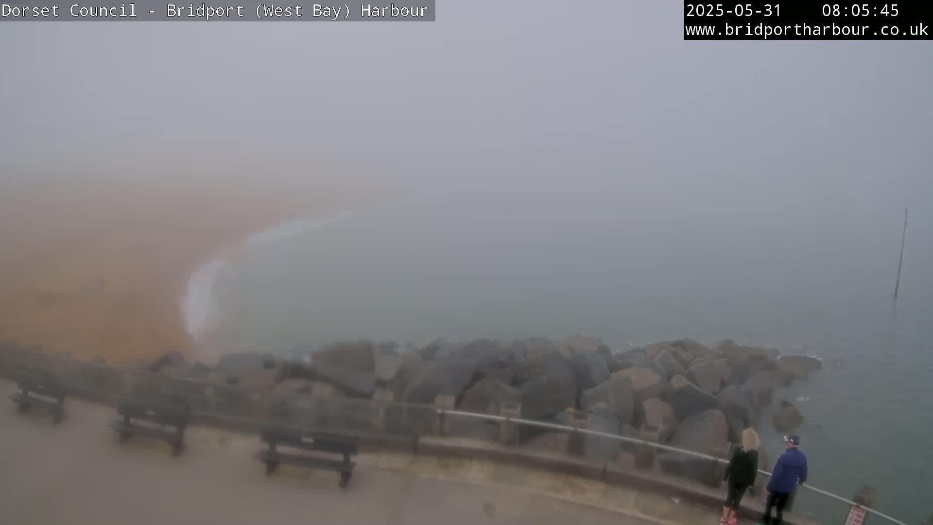 A foggy coastal scene shows a rocky breakwater, a sandy beach partially visible through the fog, and two people standing near a railing.