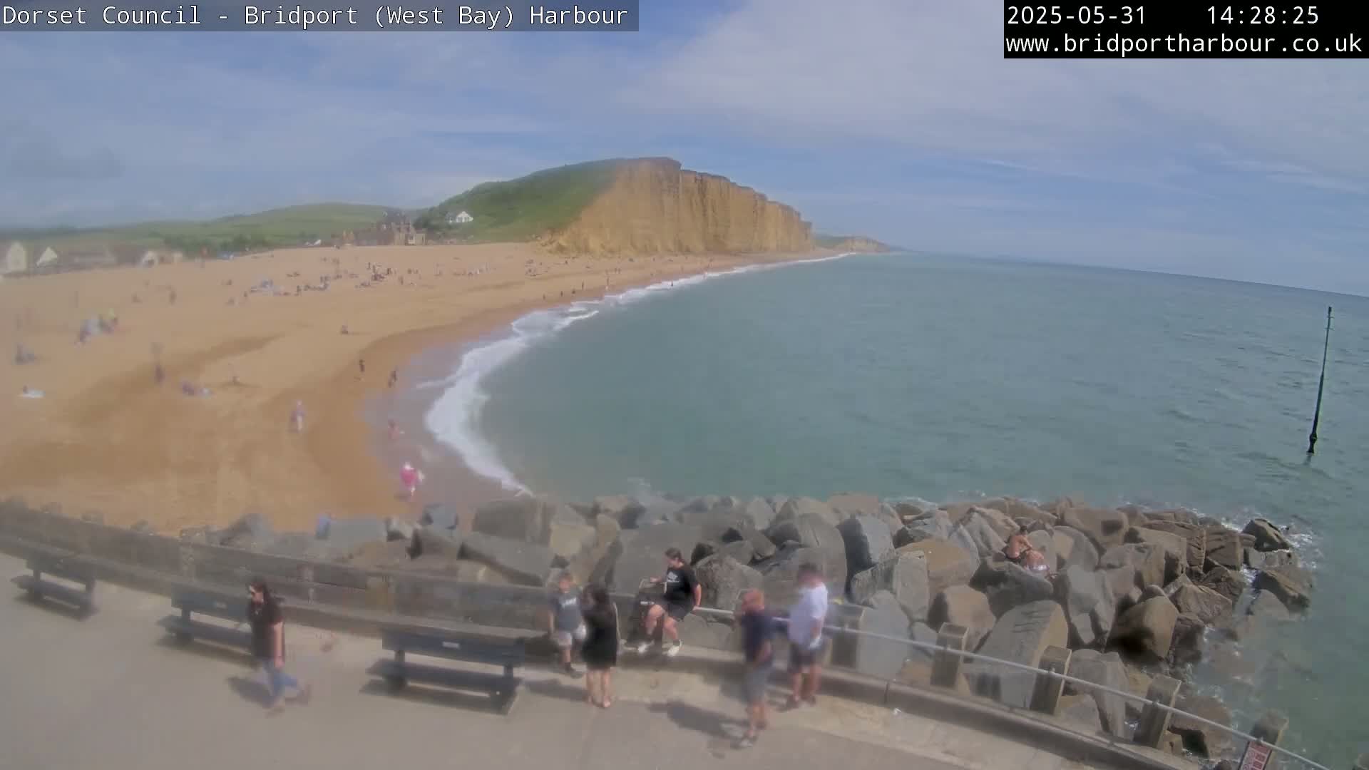 A sandy beach with many people on it curves around a rocky breakwater, meeting a calm sea under a partly sunny sky.