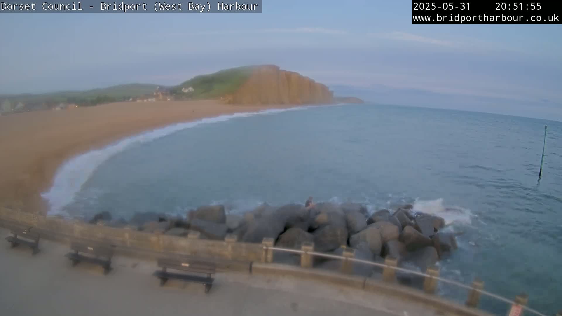 A sandy beach curves into calm, blue-green water lapping against a rocky breakwater under a mostly clear evening sky.