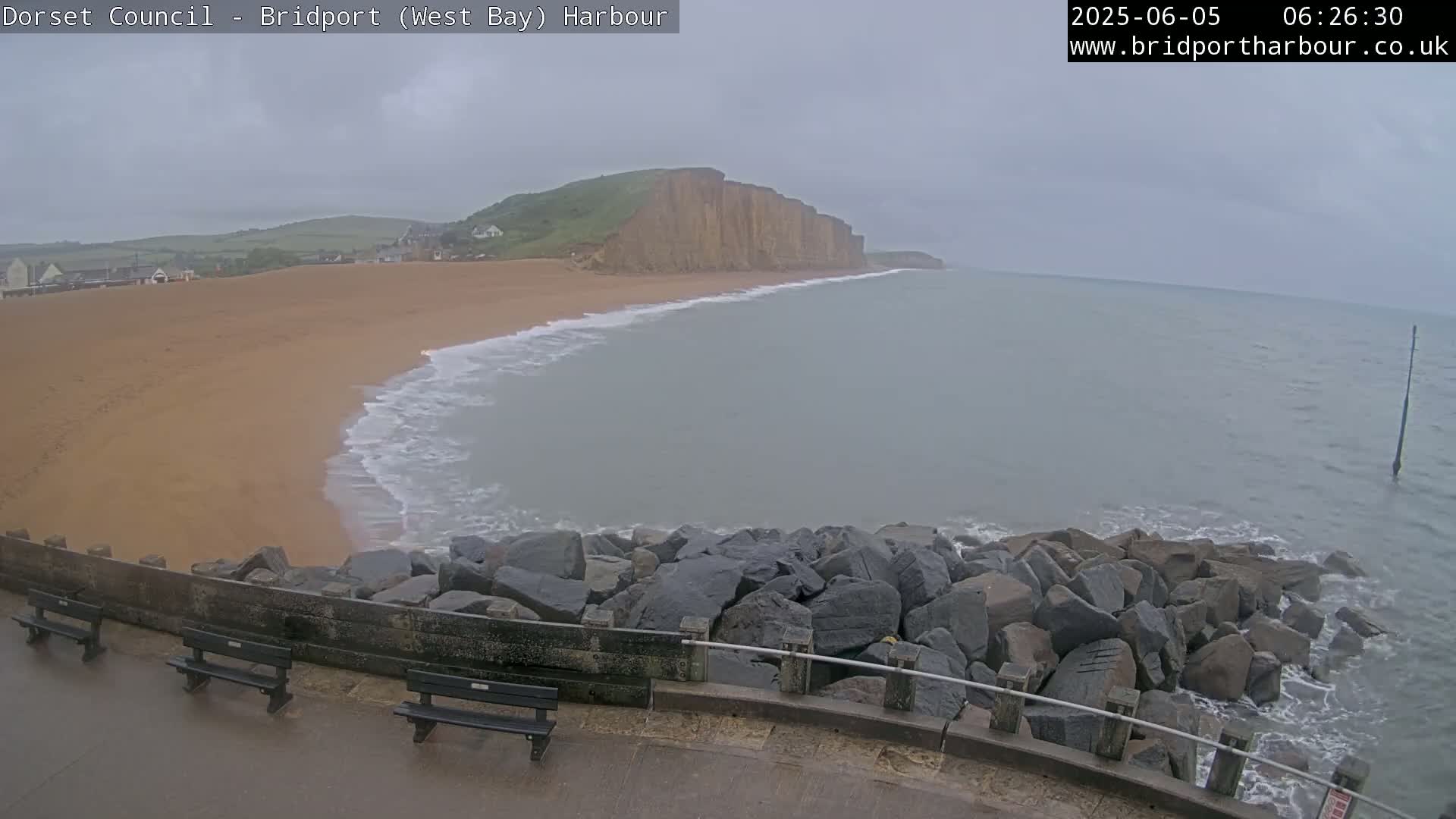 A sandy beach meets a calm, gray sea under an overcast sky, backed by a large cliff face.