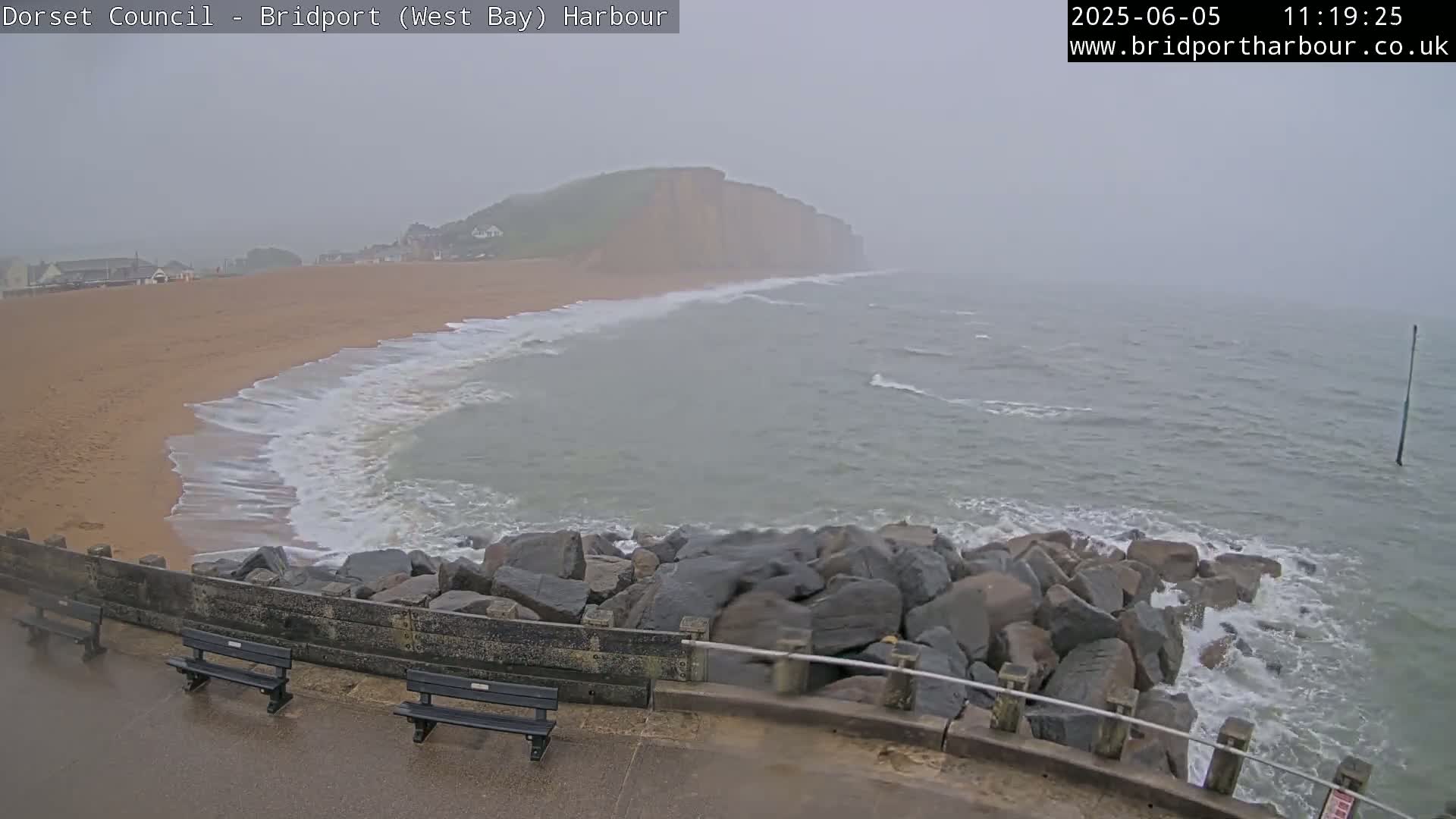 A sandy beach with moderate waves crashing against a rocky seawall under overcast skies.