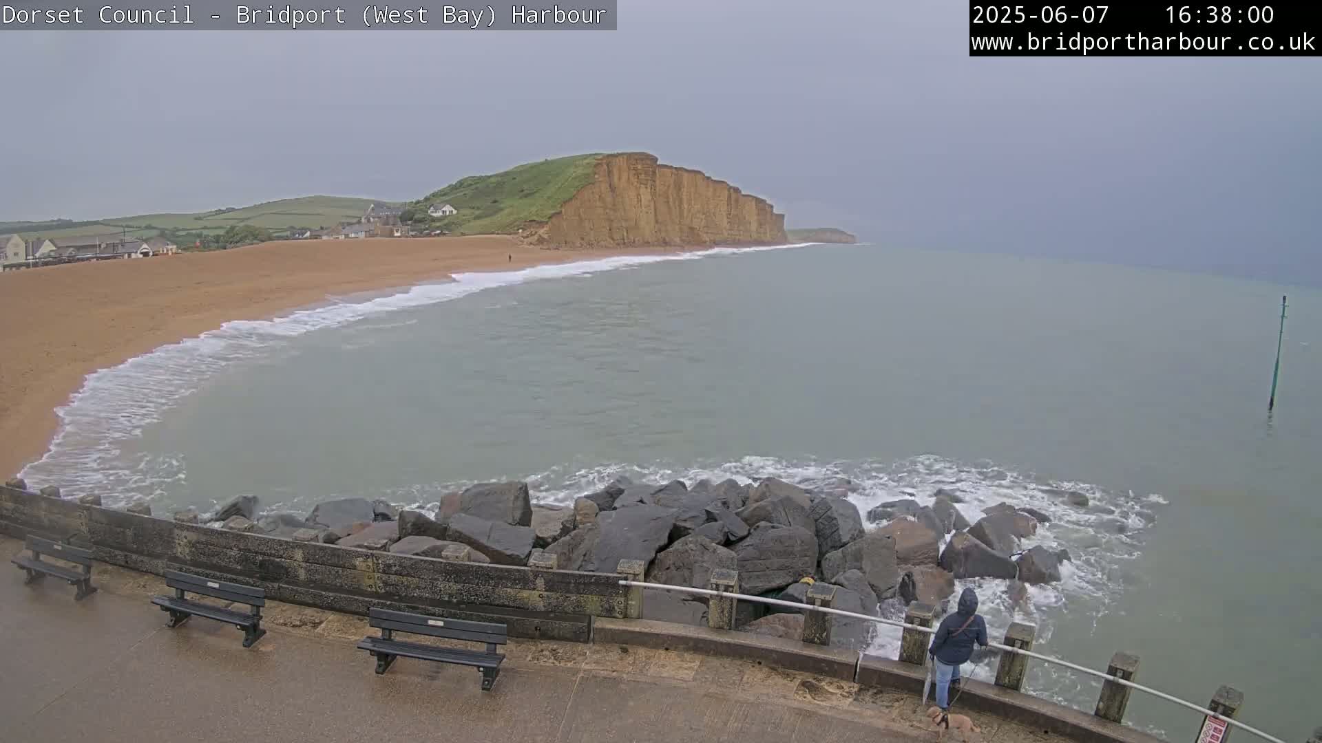 A sandy beach with calm waves laps against a rocky seawall, overlooking a grey, overcast ocean and a large cliff face in the distance.