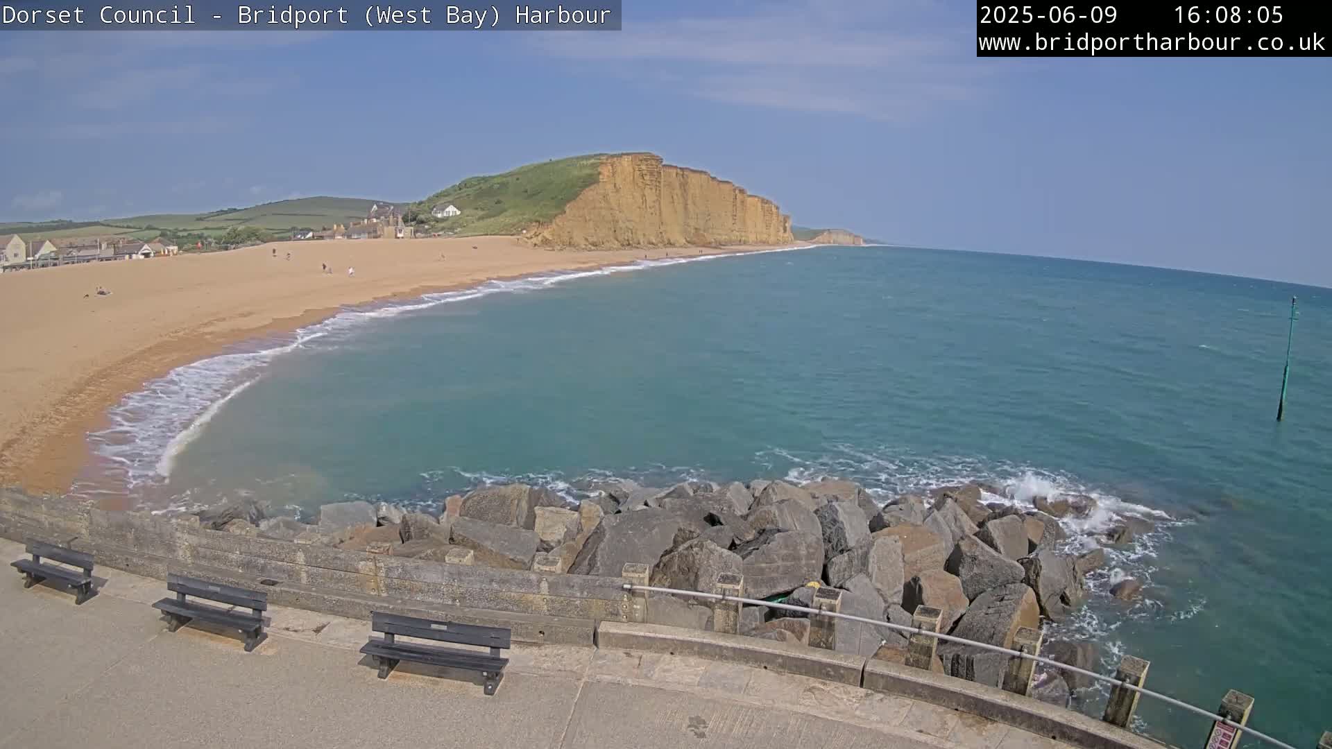 A sandy beach with a few people on it curves around a rocky outcropping into a teal ocean under a clear blue sky.