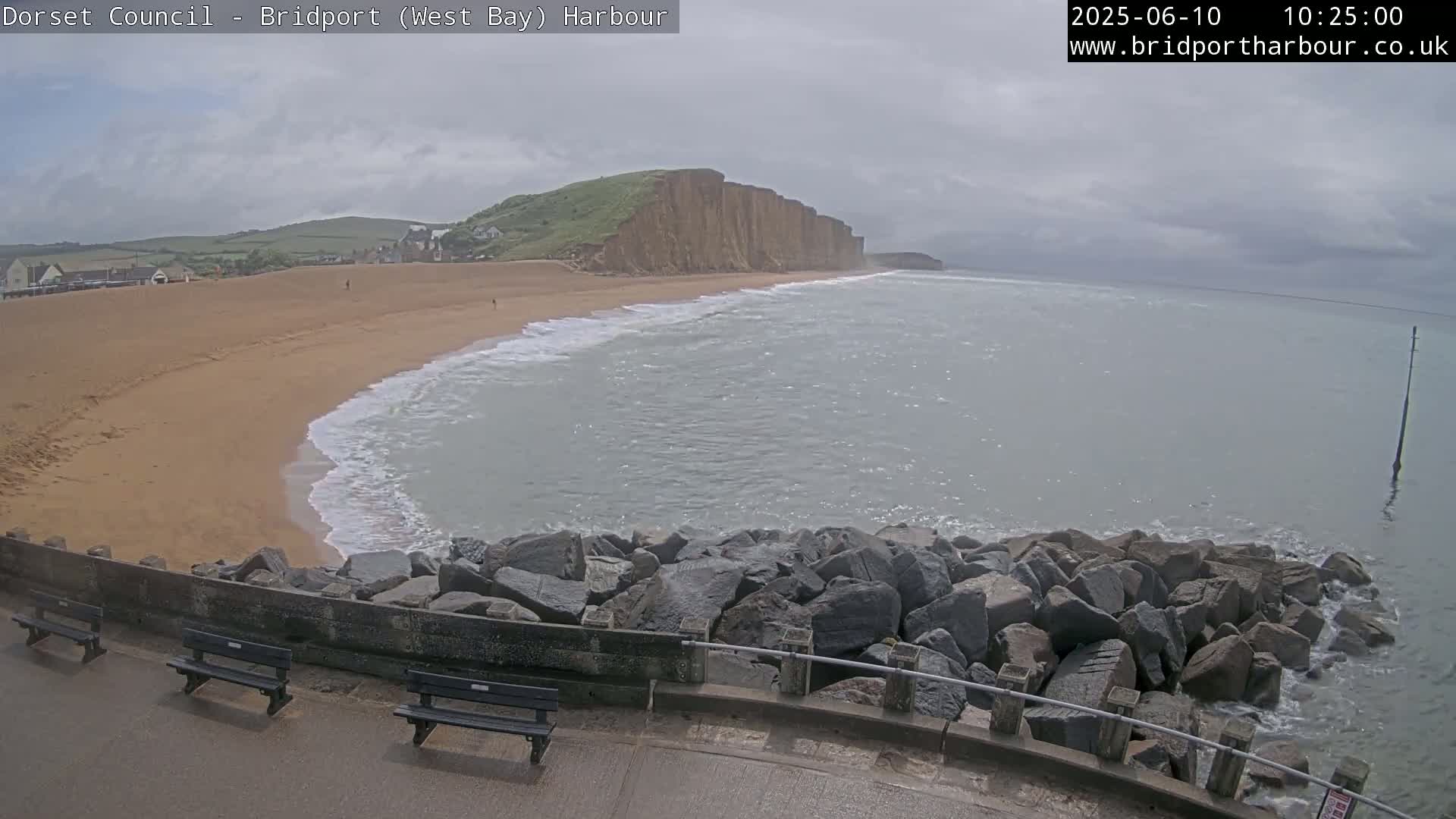 A sandy beach stretches beneath a cloudy sky, meeting a calm sea lapping against a rocky breakwater in the foreground.
