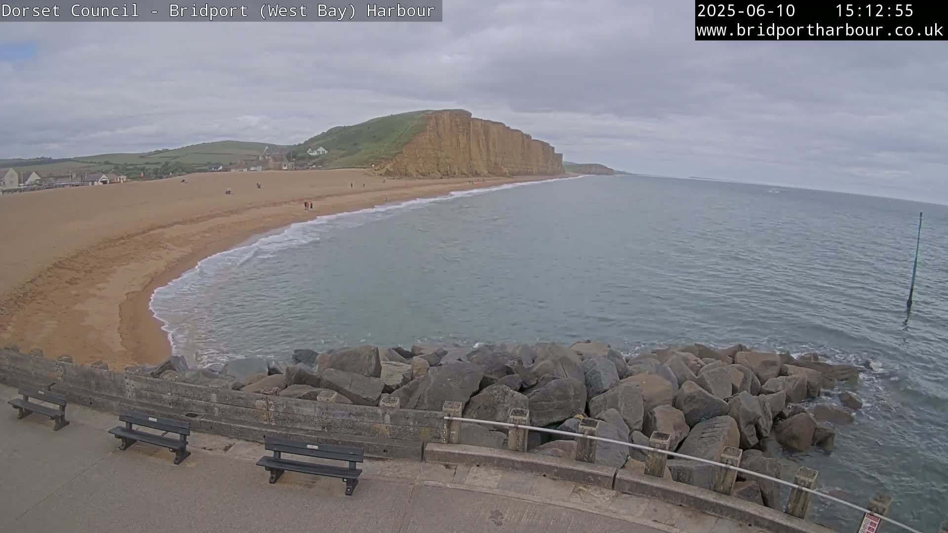 A sandy beach with a few people on it stretches out to a calm sea meeting a large cliff face under an overcast sky.