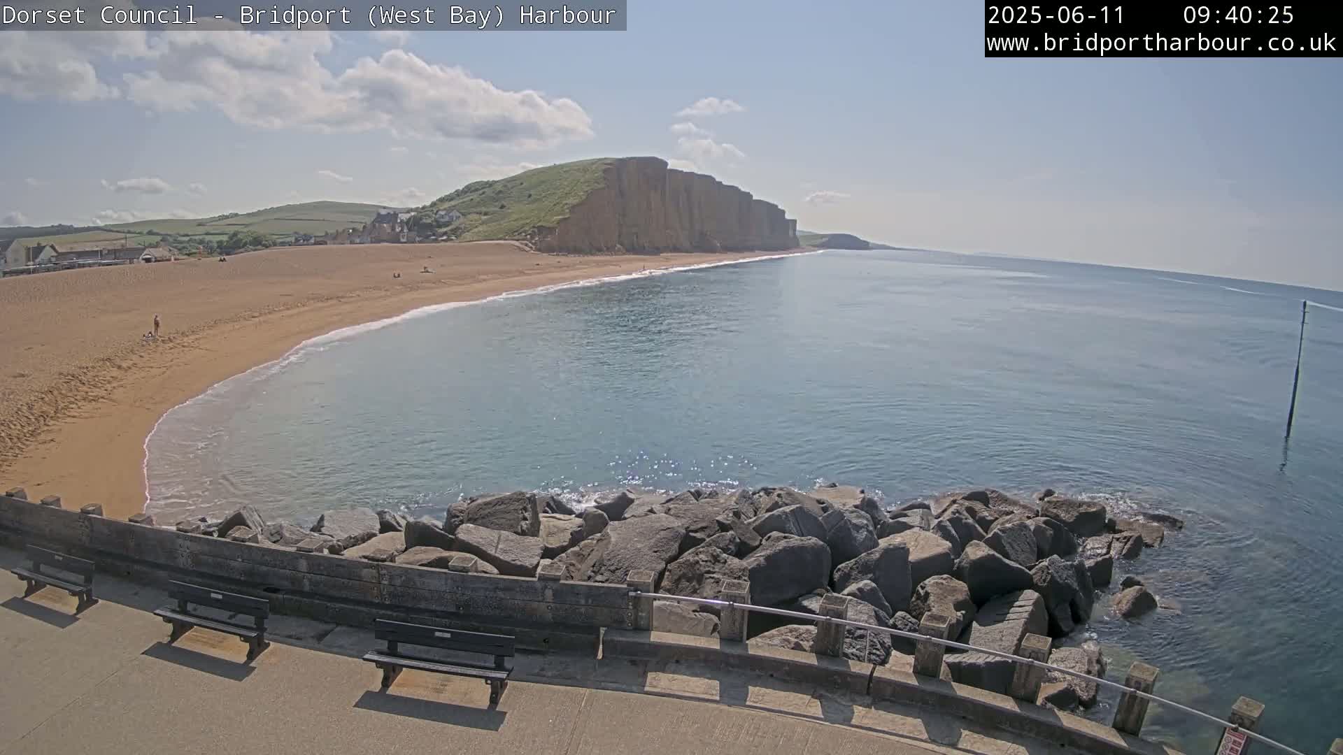 A sandy beach curves into a calm, blue ocean, backed by a high, green cliff under a partly cloudy sky.
