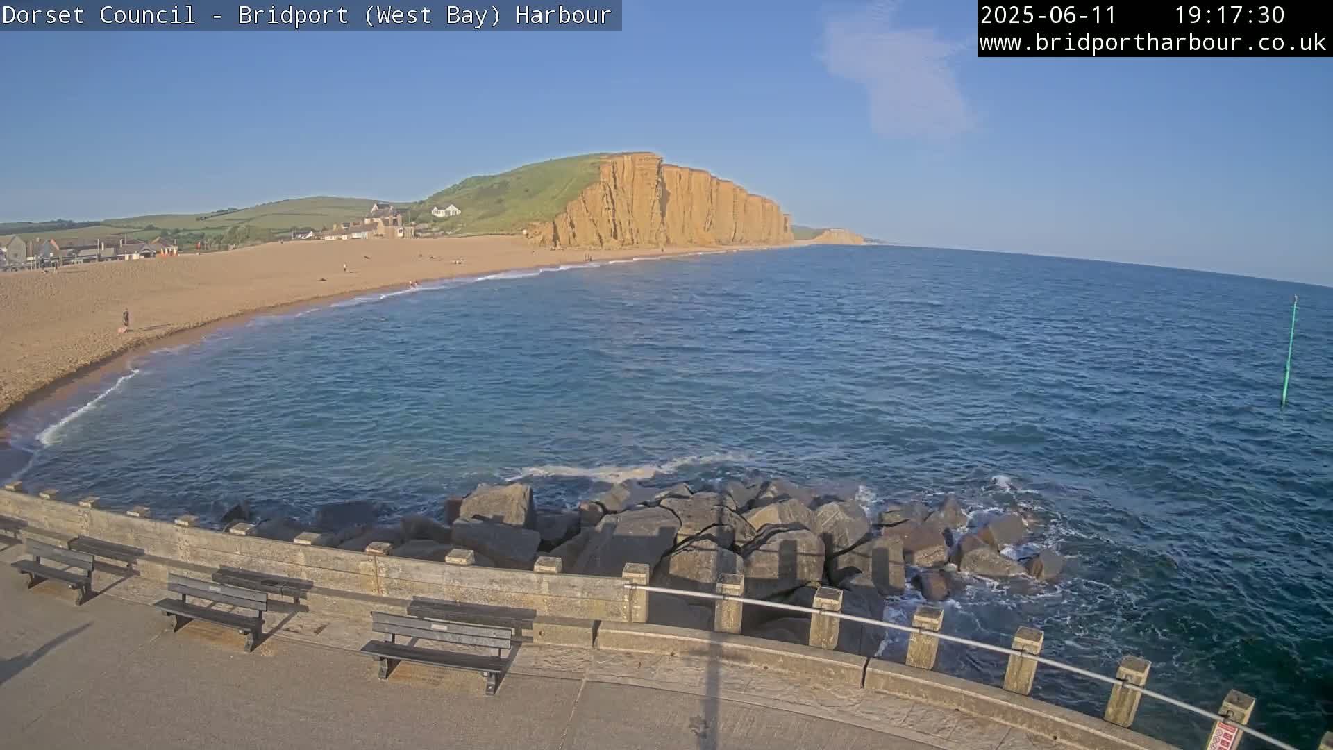 A sandy beach curves into a calm, blue ocean under a clear, sunny sky, with large cliffs visible in the distance and several benches lining a stone seawall in the foreground.