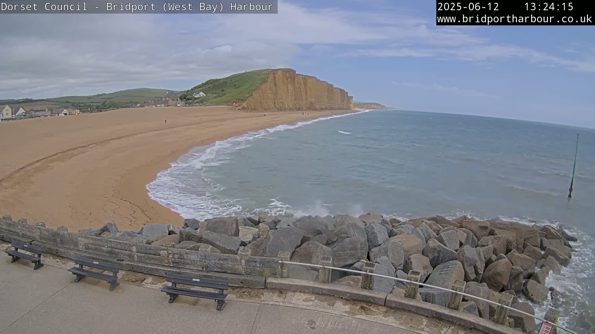 A sandy beach curves around a rocky breakwater, with a large cliff face in the background under a partly cloudy sky, and small waves lapping the shore.