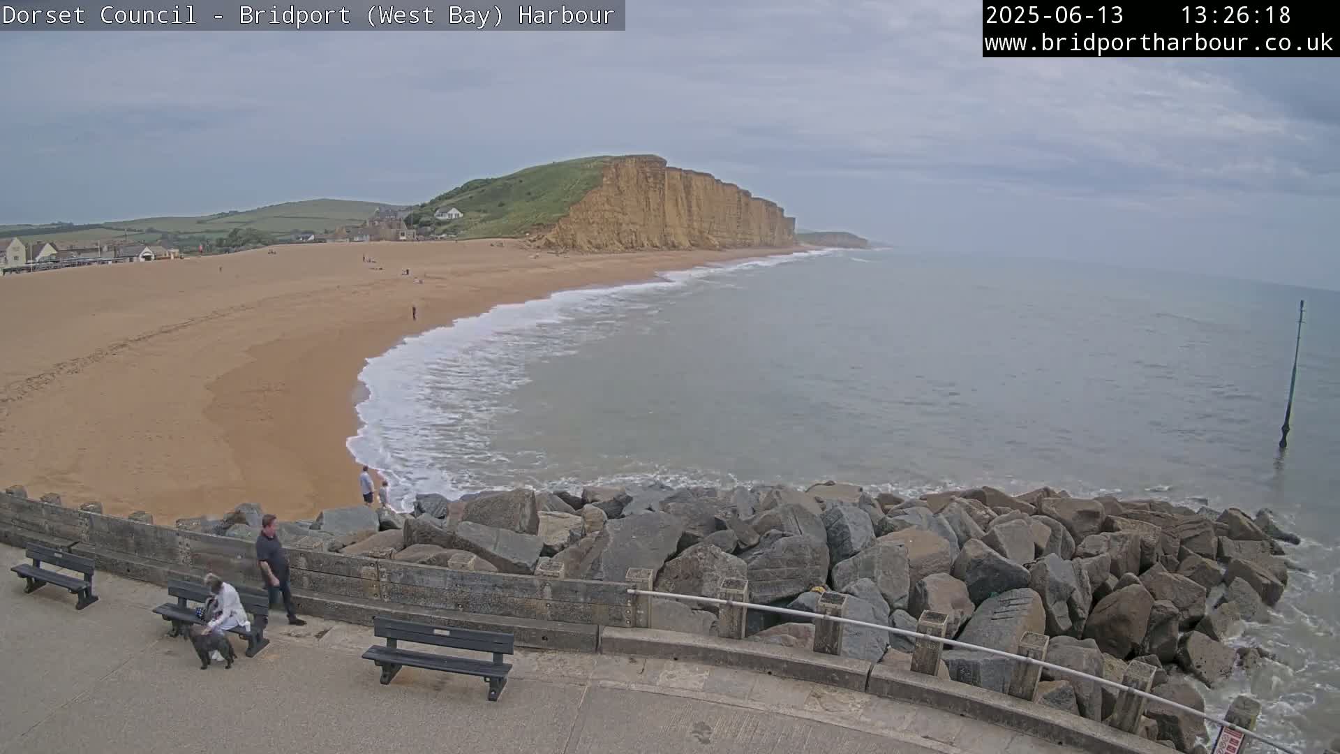 A sandy beach with a few people on it curves around a rocky headland under an overcast sky, with gentle waves lapping at the shore.