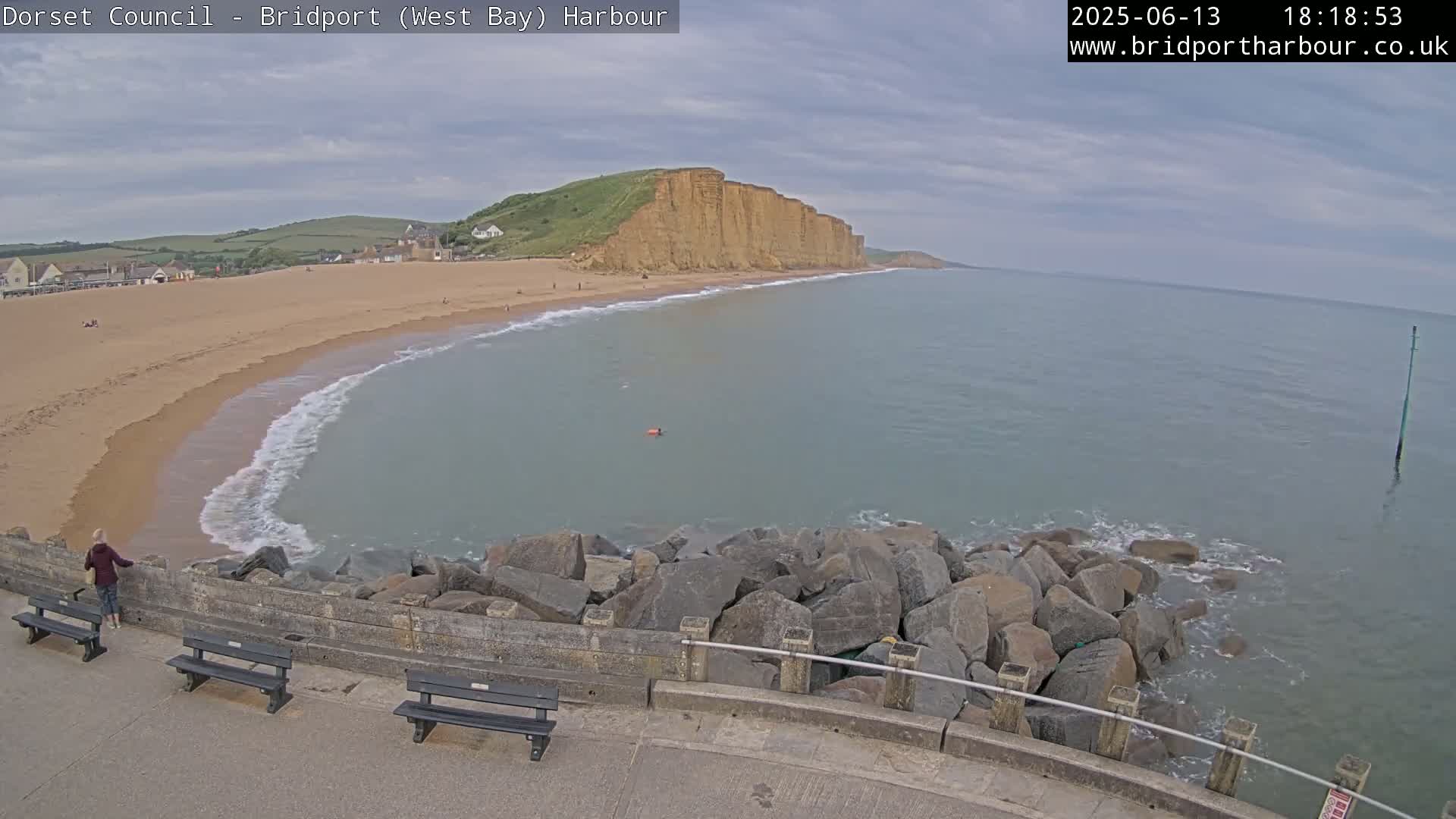 A sandy beach curves around a calm bay bordered by a rocky breakwater and high, tan cliffs under a mostly cloudy sky.
